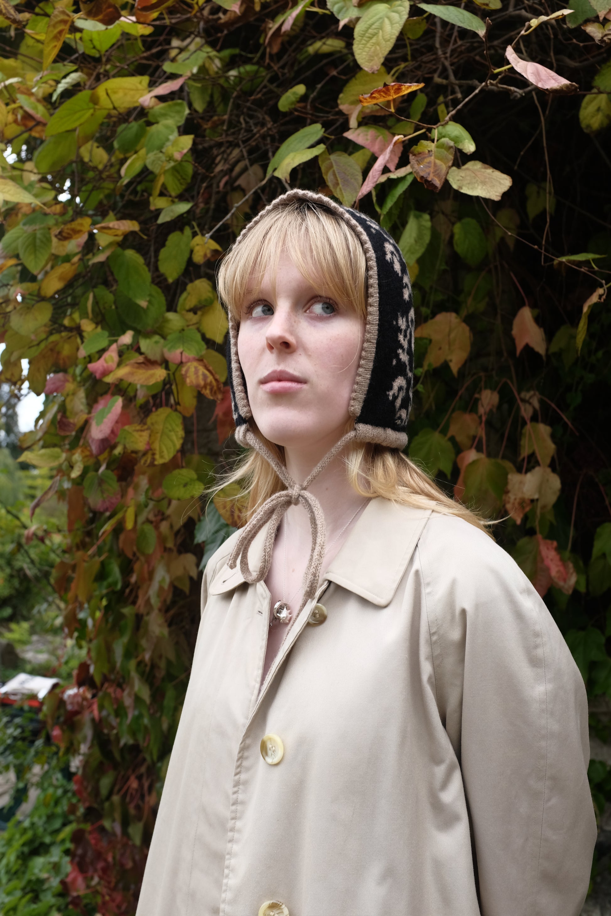 A side angled view of a women weaering a cashmere and lambswool knitted bonnet. The Bonnets base is black whilst the trim, pattern and tie are tan. The pattern on the bonnet consists of leaves and mushrooms sequenced satisfyingly up the side of the hood. Behind the women is a wave of green, red and orange leaves. 