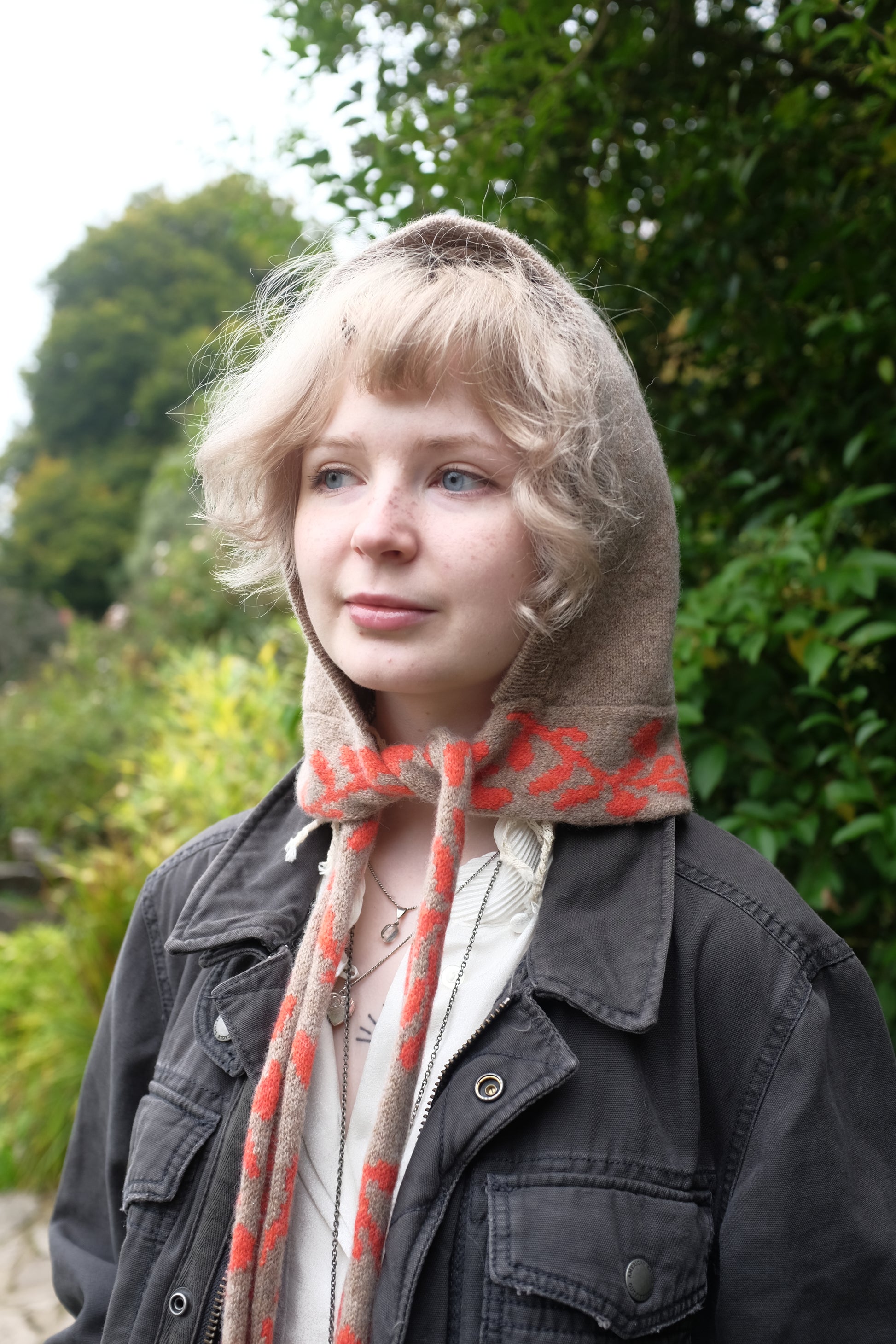 A front angled view of a woman wearing a lambswool knitted bonnet hood. The base of the bonnet is a tan colour. There is a fire orange pattern on the bottom of the bonnet tie that vis longer, mimicking a scarf. The pattern on the is a sequenced of laurel leaves and hearts. The women has tied the scarf loosely and behind her is a mix of green autumnal leaves 