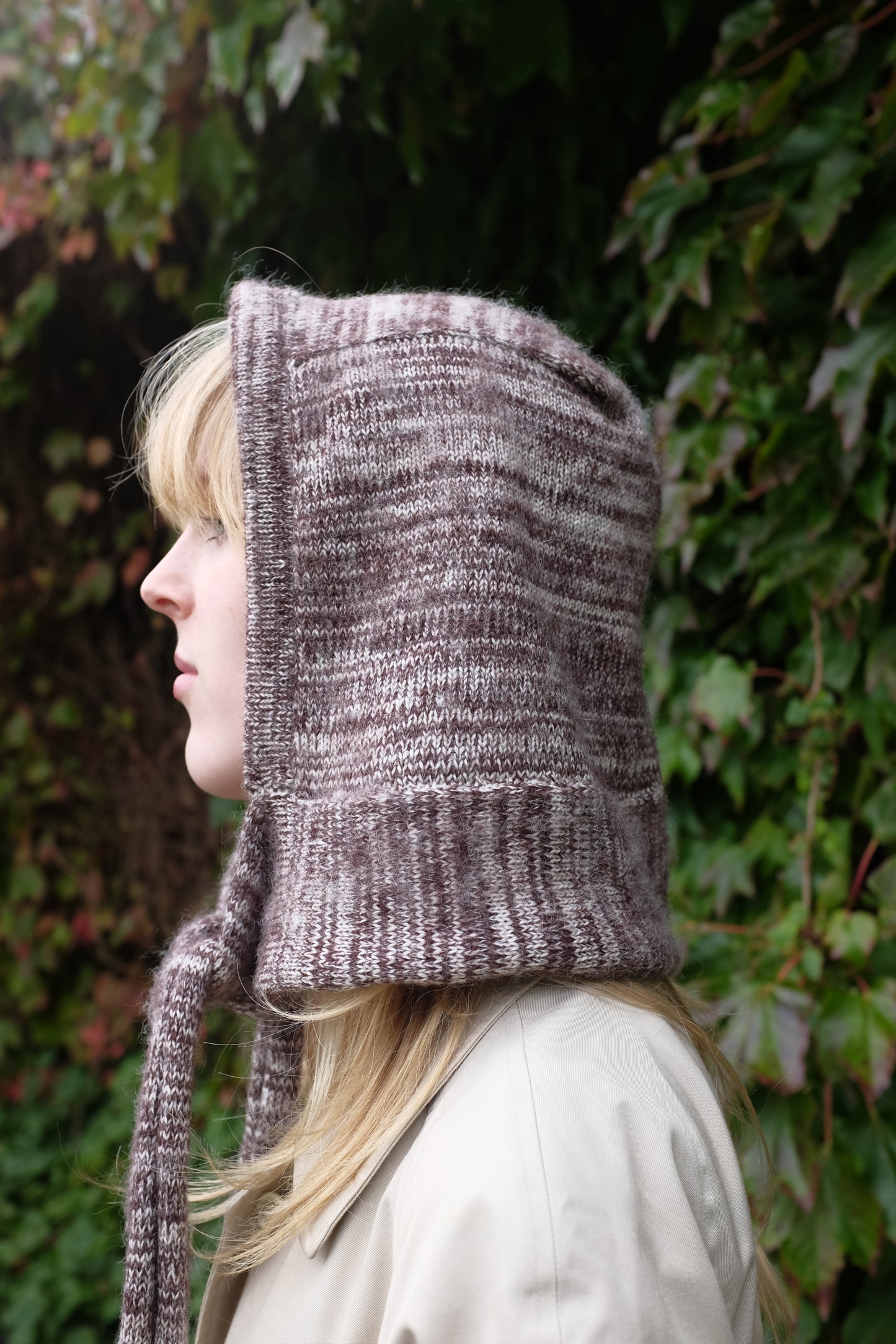 Person wearing a burgundy mohair bonnet-style hood that fully covers the head and neck. With two long, soft extensions hanging down from the front. The hood is thick and textured, offering warmth and a cosy appearance, ideal for cold weather. The person is dressed in a light coloured coat and stands outdoors against a backdrop of green and red foliage 