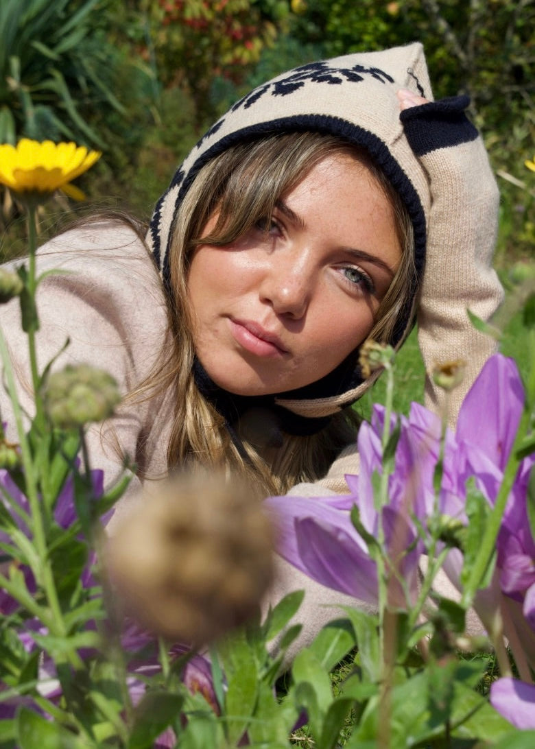 Woman lying among flowers wearing a patterned bonnet in navy and cresam and light-colored sweater.