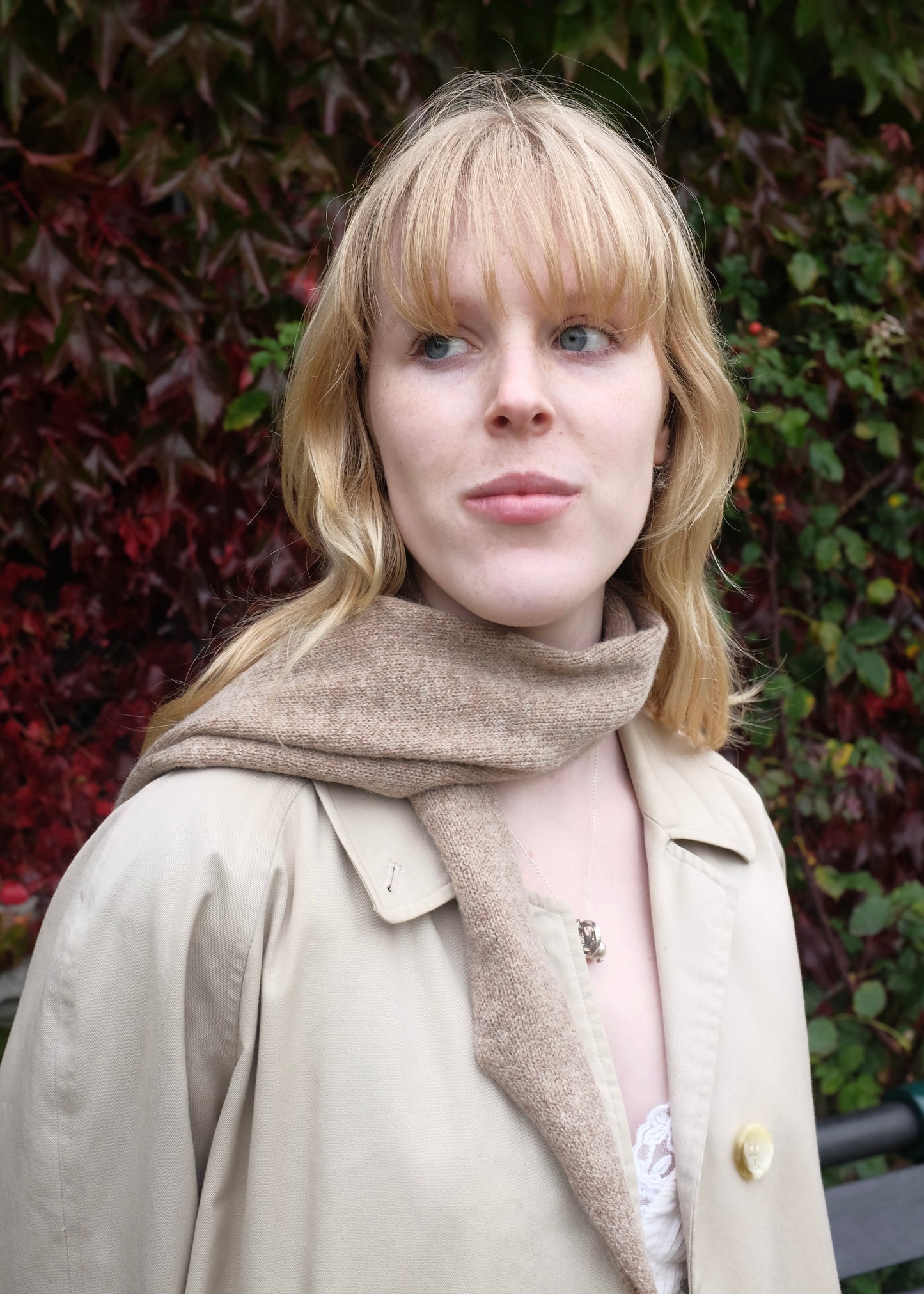 Woman wearing a beige coat and scarf against a leafy background