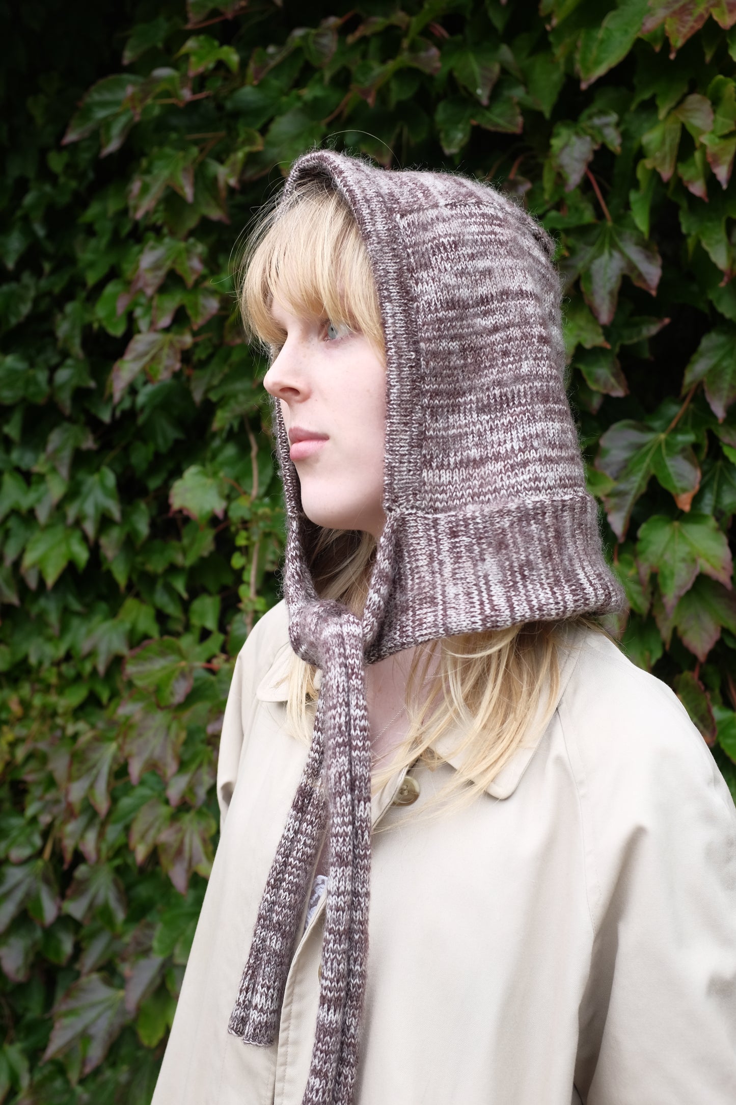 Person wearing a burgundy mohair bonnet-style hood that fully covers the head and neck. With two long, soft extensions hanging down from the front. The hood is thick and textured, offering warmth and a cosy appearance, ideal for cold weather. The person is dressed in a light coloured coat and stands outdoors against a backdrop of green and red foliage 