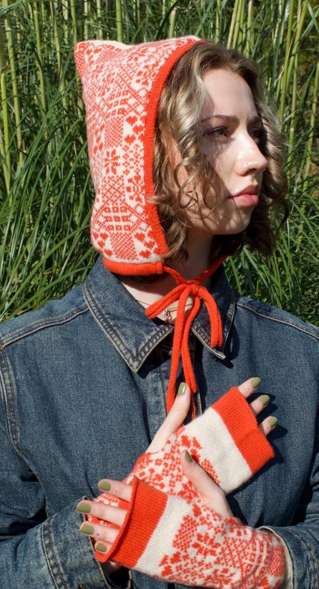 A person is outdoors side view , surrounded by tall grass and tress, wearing a hand-knitted cashmere hood bonnet. The hood features a symmetrical orange and white pattern inspired by traditional Nordic or Fair isle designs. It ties neatly under thr chin with an orange string, showcasing detailed craftsmanship and cultural textile artistry. 
