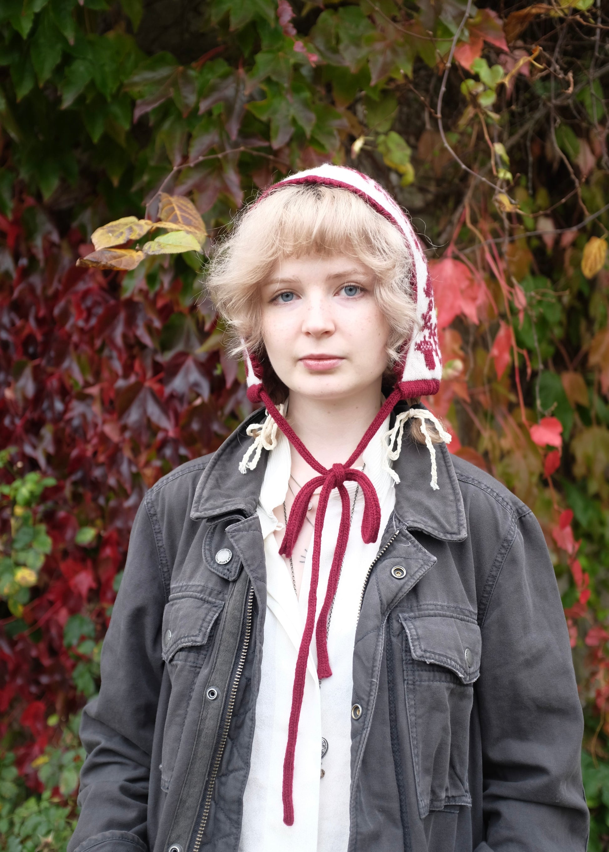 Person wearing a knitted cream and burgundy bonnet with a bow in front of leafy green and red background
