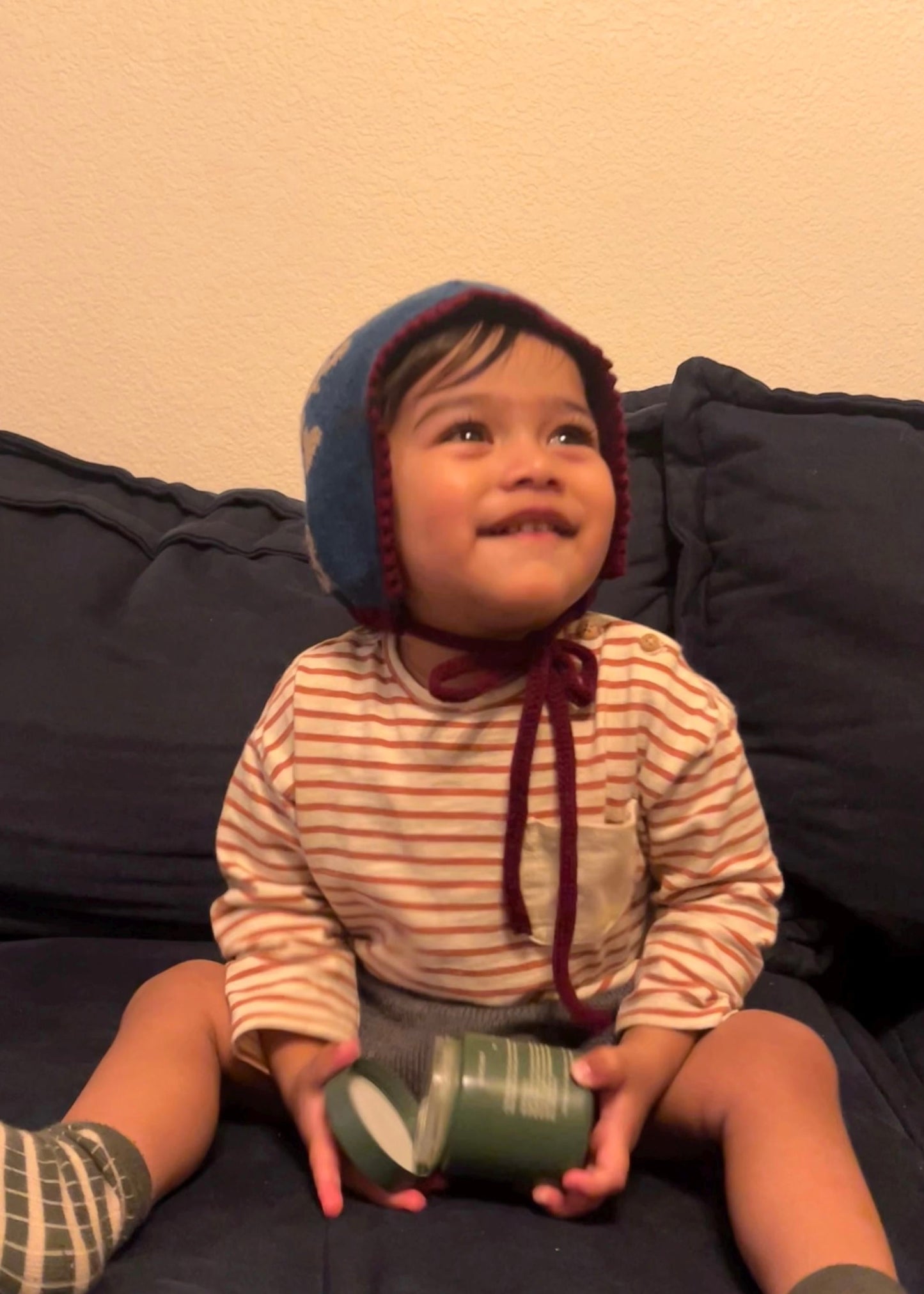 Child wearing a striped shirt and blue, holding a green toy, sitting on a dark surface with a beige background.