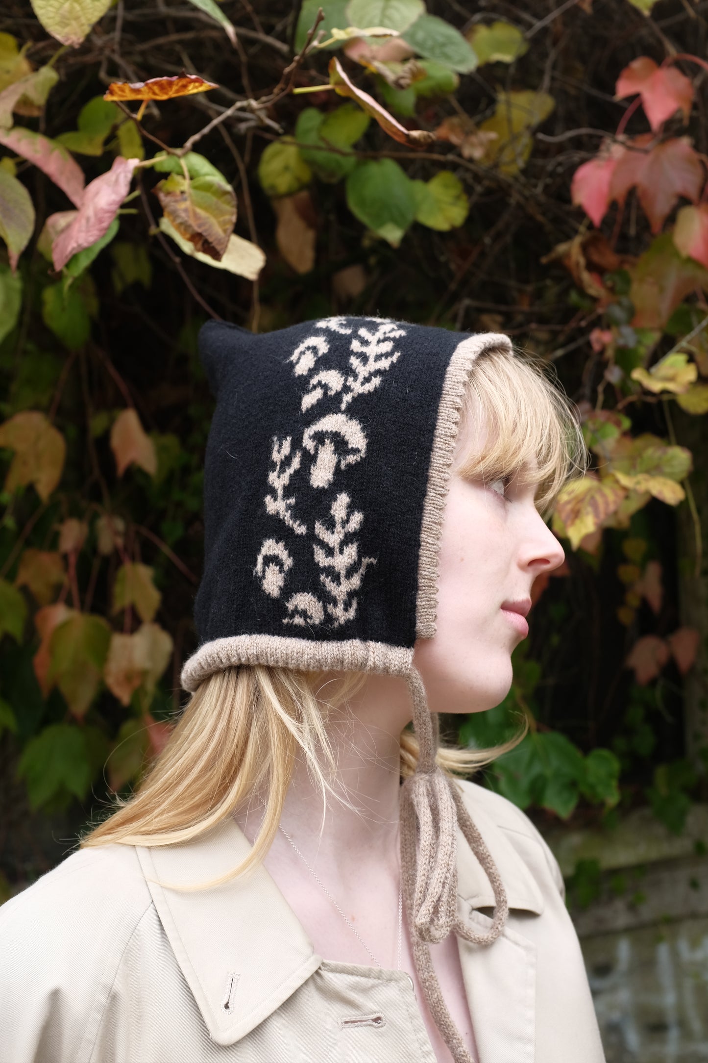 A side view of a women weaering a cashmere and lambswool knitted bonnet. The Bonnets base is black whilst the trim, pattern and tie are tan. The pattern on the bonnet consists of leaves and mushrooms sequenced satisfyingly up the side of the hood. Behind the women is a wave of green, red and orange leaves. 
