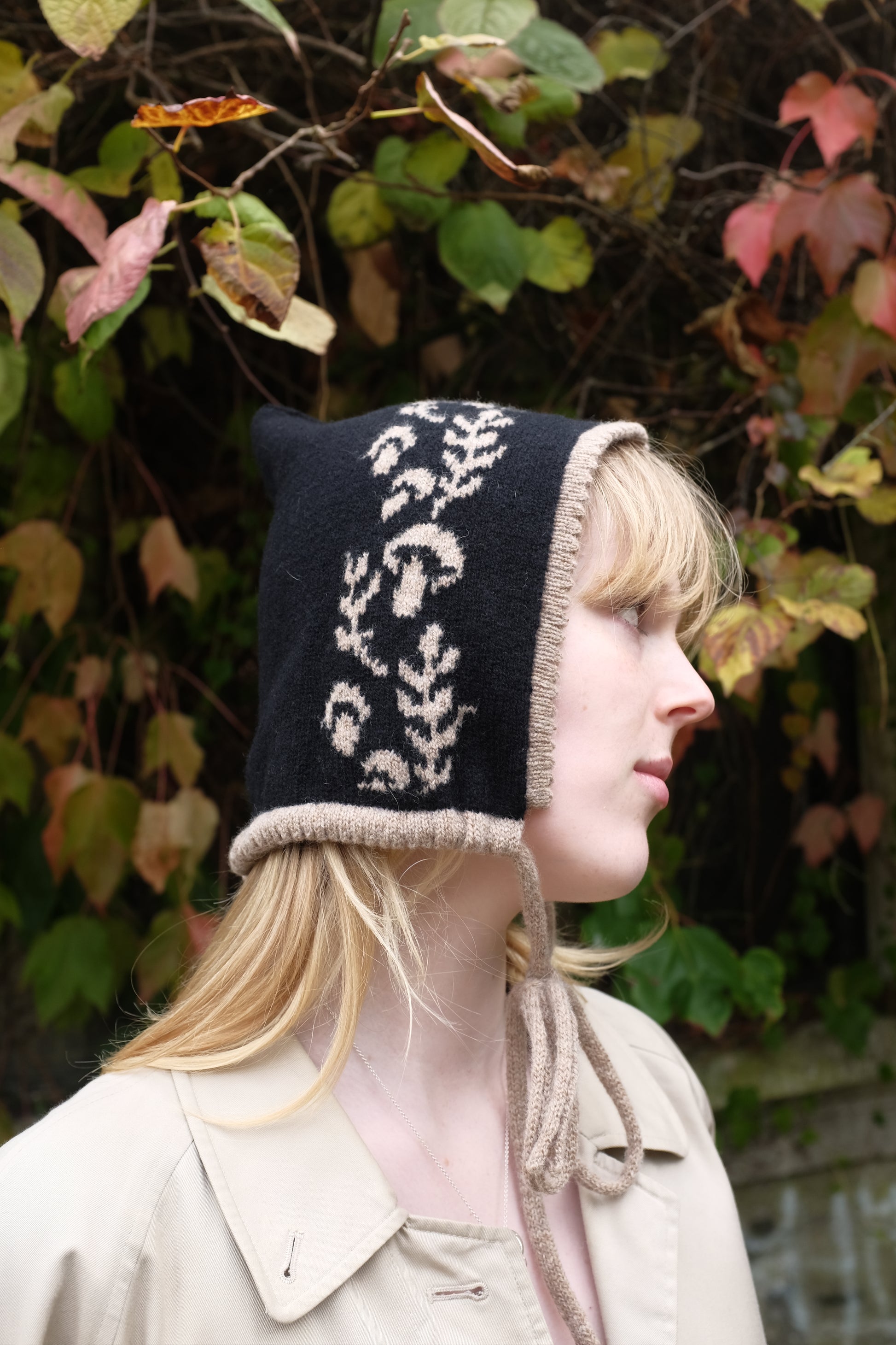 A side view of a women weaering a cashmere and lambswool knitted bonnet. The Bonnets base is black whilst the trim, pattern and tie are tan. The pattern on the bonnet consists of leaves and mushrooms sequenced satisfyingly up the side of the hood. Behind the women is a wave of green, red and orange leaves. 