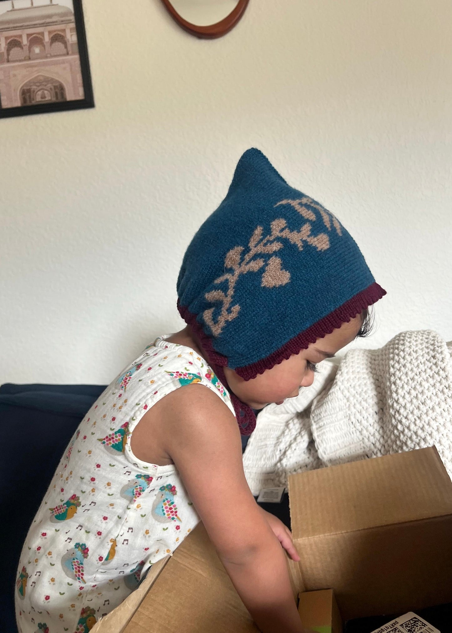 Child wearing a blue knit hat with a pattern, sitting on a couch and looking into a cardboard box.
