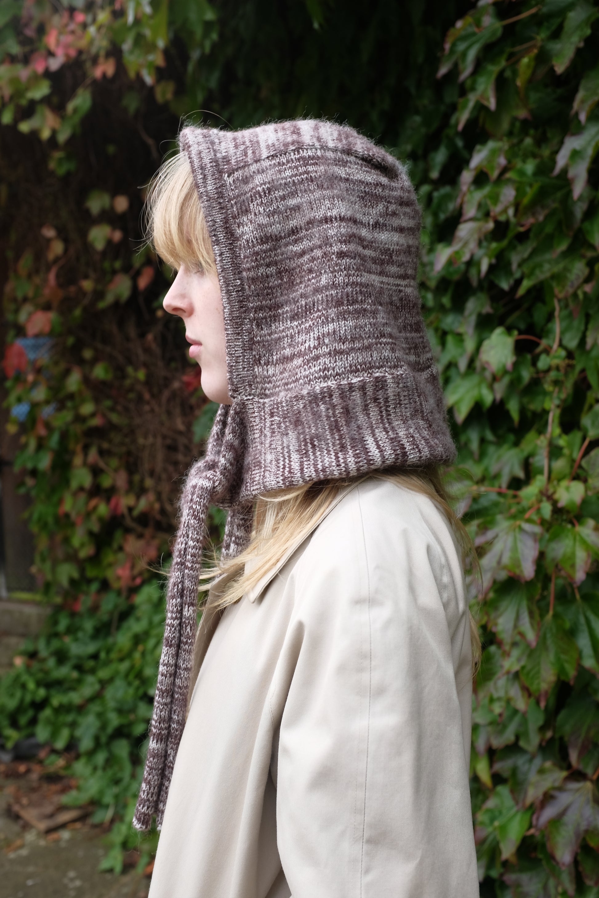 Person wearing a burgundy mohair bonnet-style hood that fully covers the head and neck. With two long, soft extensions hanging down from the front. The hood is thick and textured, offering warmth and a cosy appearance, ideal for cold weather. The person is dressed in a light coloured coat and stands outdoors against a backdrop of green and red foliage 