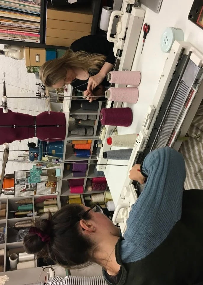 Two people working on a knit machine in a craft room with shelves and supplies.