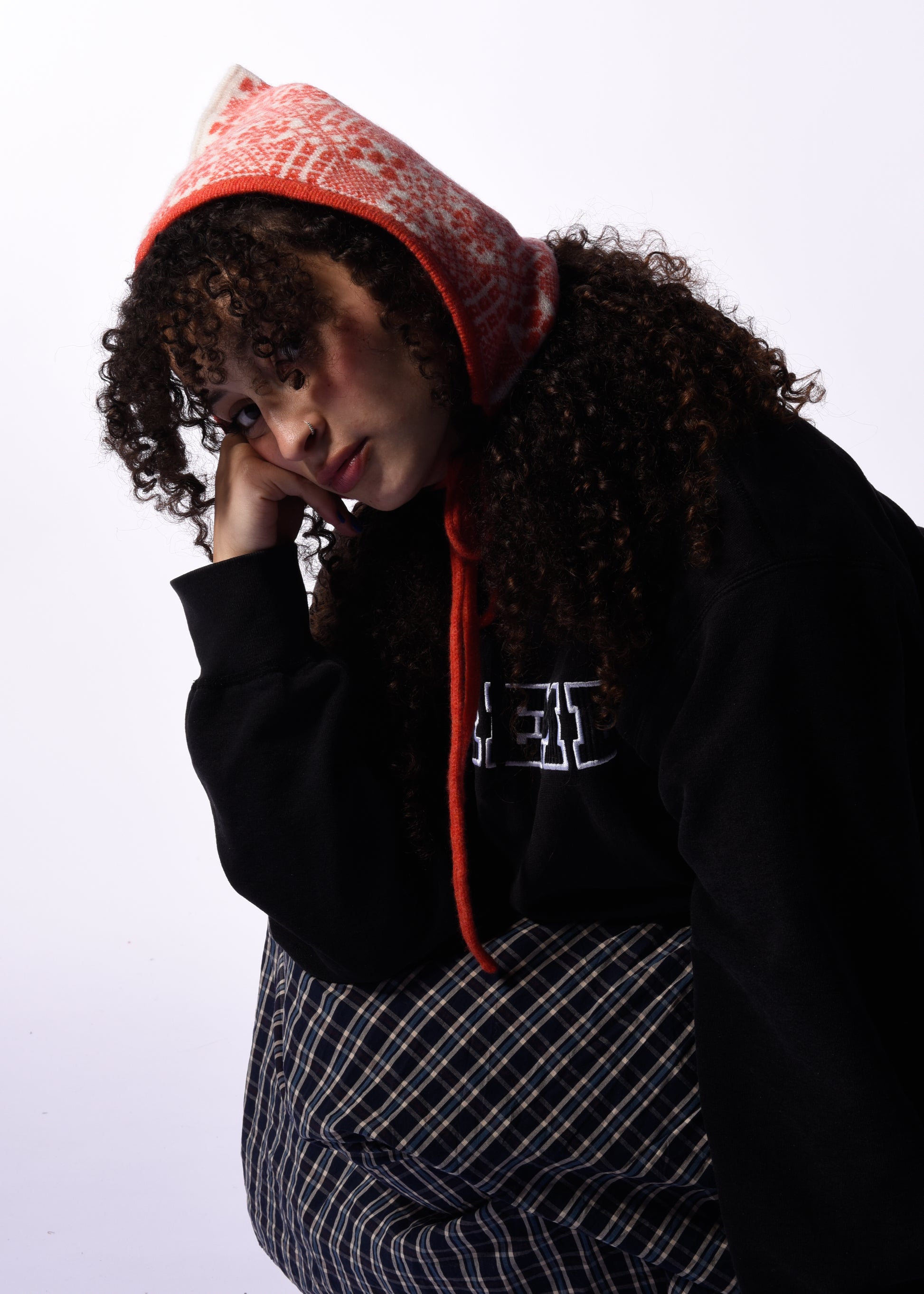 Person wearing a black hoodie with orange bonnet and plaid pants on a white background
