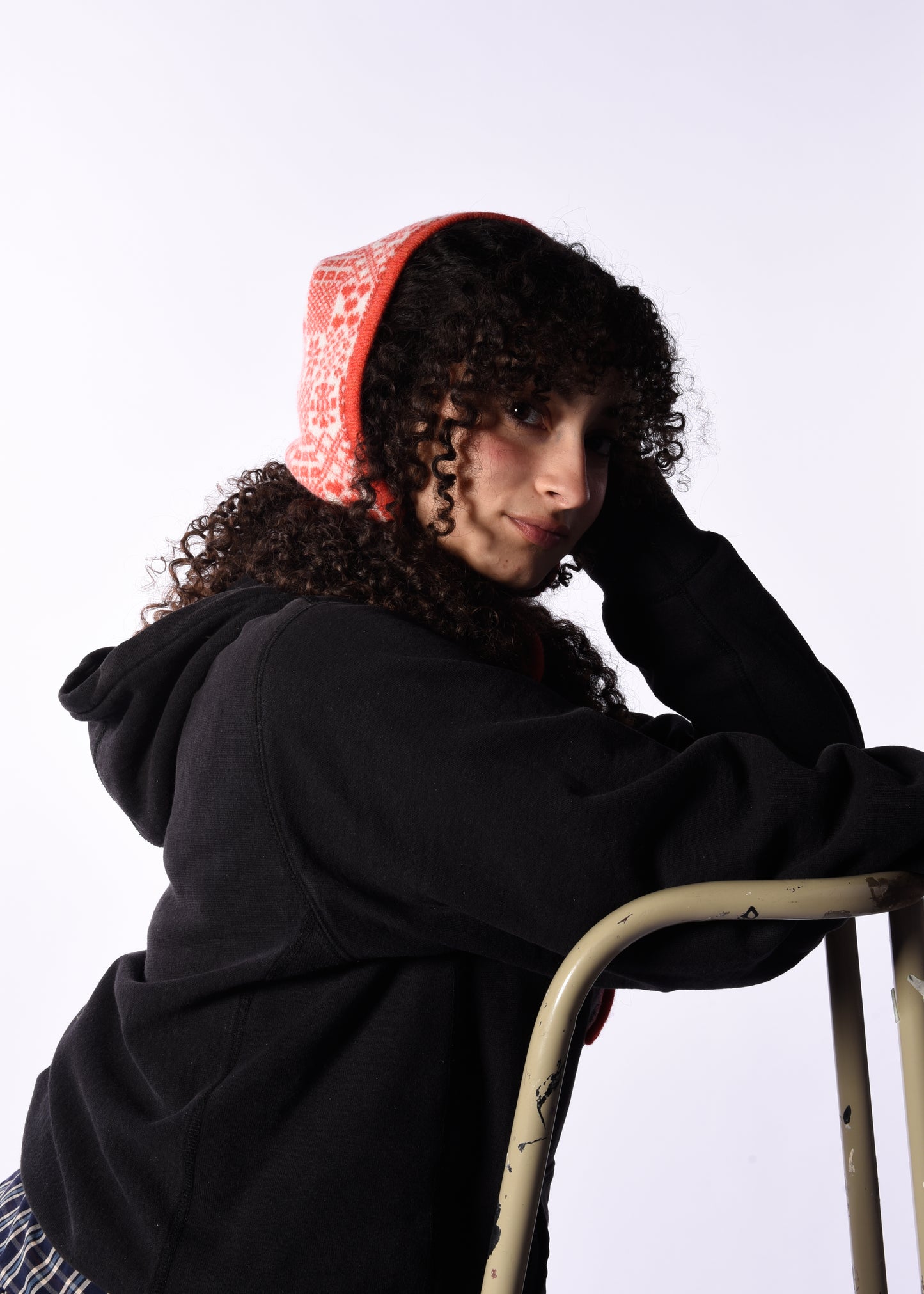 Person wearing a black hoodie with a orange hood, leaning against a white wall.