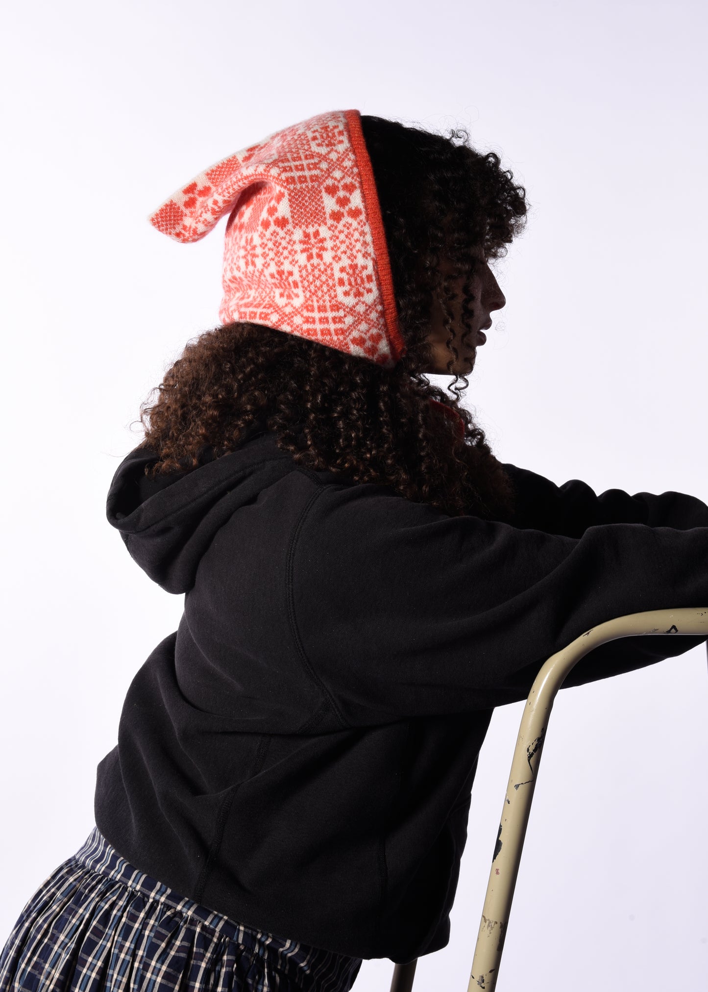 Person wearing a black hoodie and plaid skirt with a orange patterned bonnet on a white background
