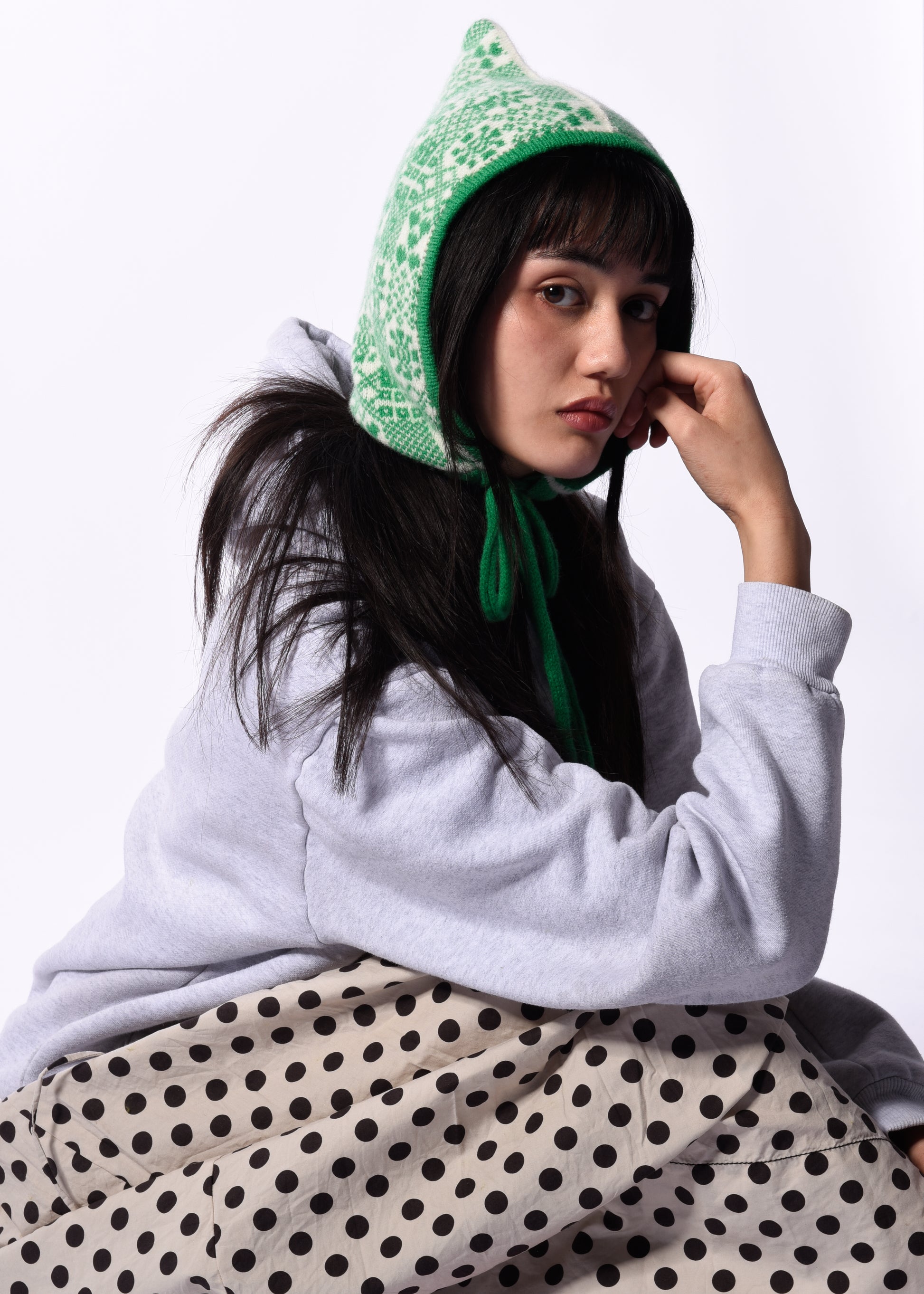 Person wearing a green patterned bonnet, gray sweater, and polka dot skirt on a white background