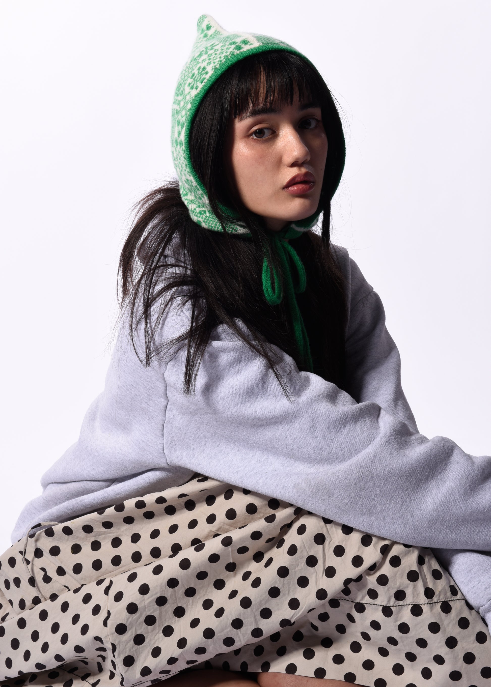 Person wearing a light gray hoodie, green bonnet, and polka dot pants on a white background