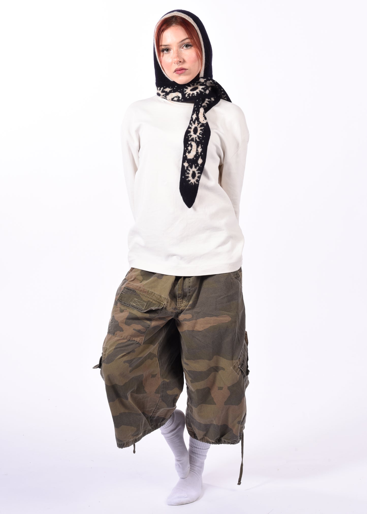 Person wearing a white hoodie, camouflage pants, and a patterned scarf bonnet on a white background
