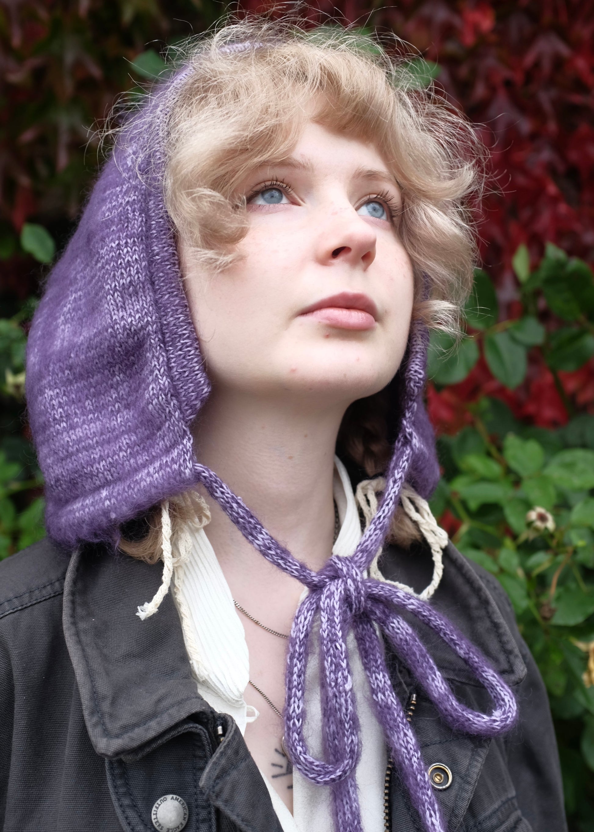 Person wearing a purple knitted bonnet against a green and red leafy background
