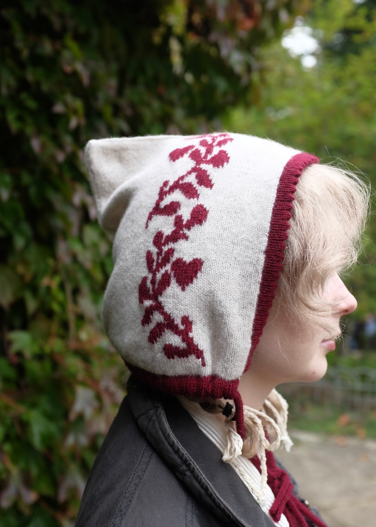Person wearing a white knitted bonnet with red laurel leaf design, trim and tie outdoors.