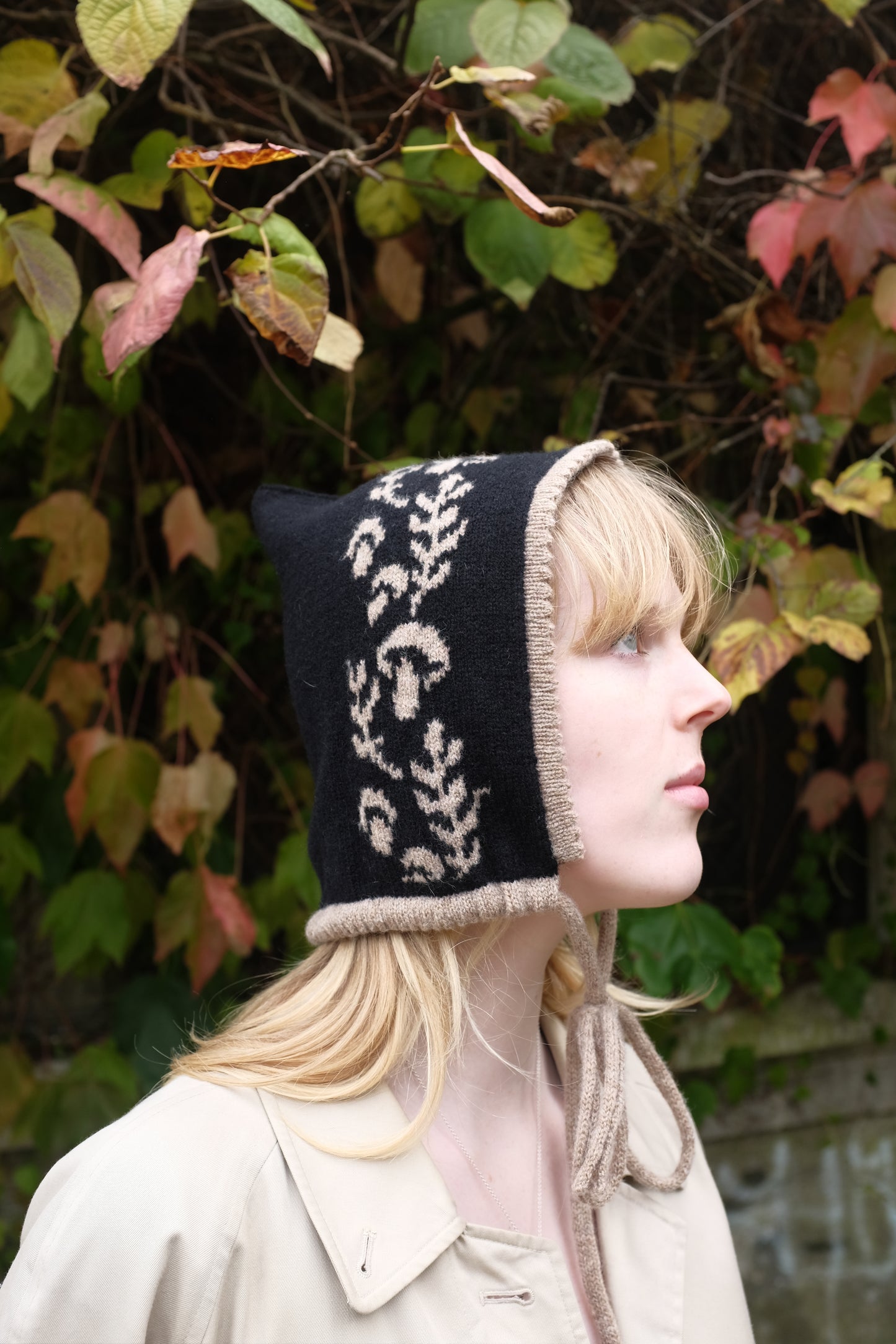 A side view of a women weaering a cashmere and lambswool knitted bonnet. The Bonnets base is black whilst the trim, pattern and tie are tan. The pattern on the bonnet consists of leaves and mushrooms sequenced satisfyingly up the side of the hood. Behind the women is a wave of green, red and orange leaves. 