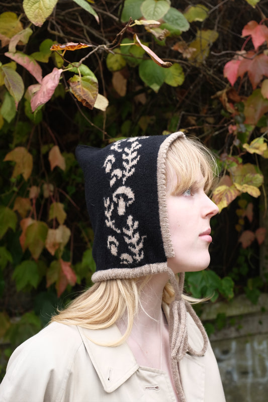 A side view of a women weaering a cashmere and lambswool knitted bonnet. The Bonnets base is black whilst the trim, pattern and tie are tan. The pattern on the bonnet consists of leaves and mushrooms sequenced satisfyingly up the side of the hood. Behind the women is a wave of green, red and orange leaves. 