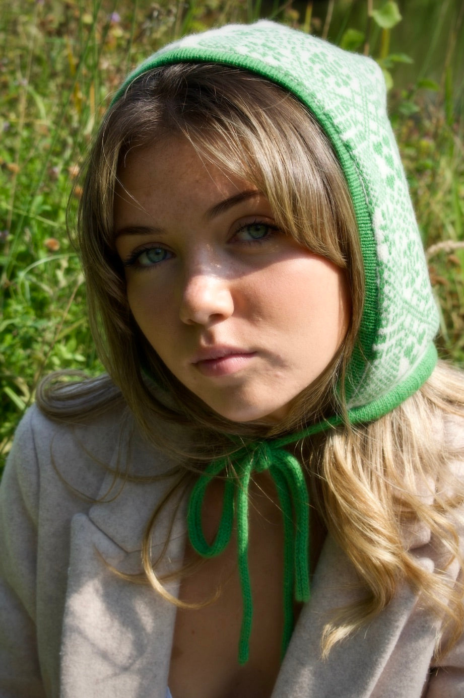 A person is outdoors, surrounded by tall grass and tress, wearing a hand-knitted cashmere hood bonnet. The hood features a symmetrical green and white pattern inspired by traditional Nordic or Fair isle designs. It ties neatly under thr chin with a green string, showcasing detailed craftsmanship and cultural textile artistry. 