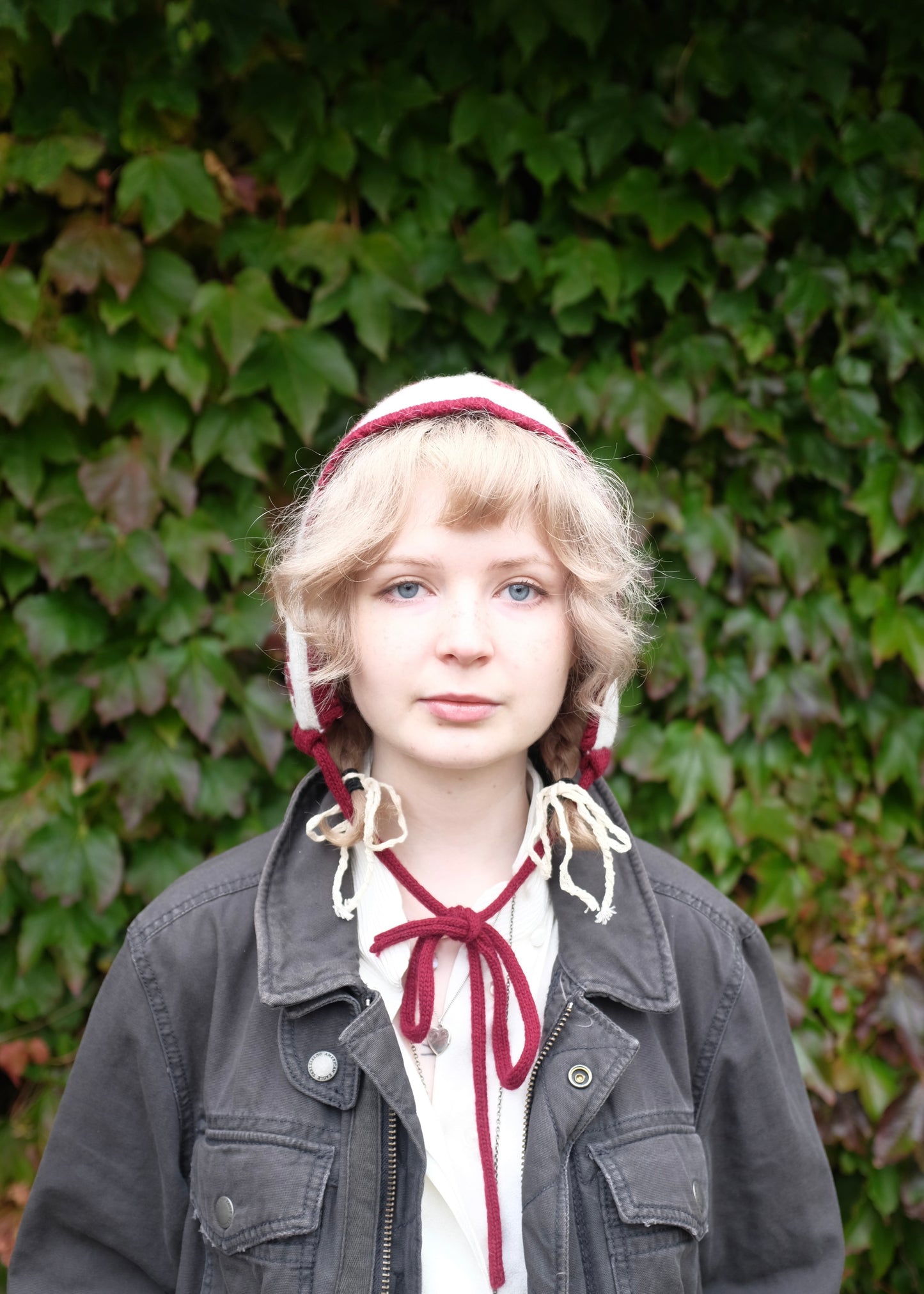 Person wearing our cashmere knitted red laurel leaf. Bonnet with a cream base, standing against a green ivy-covered wall.