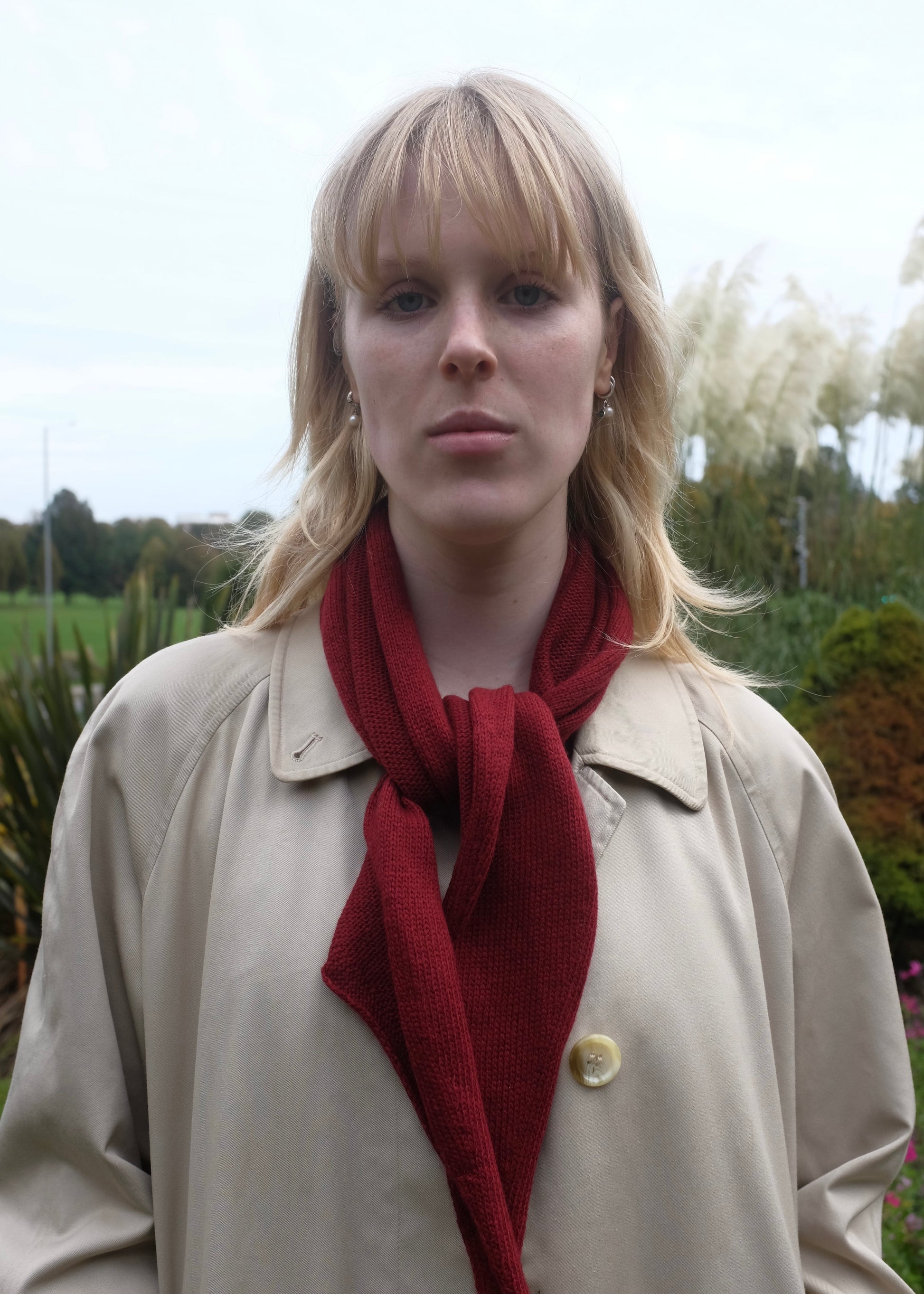 Woman wearing a beige coat and red scarf outdoors with trees in the background