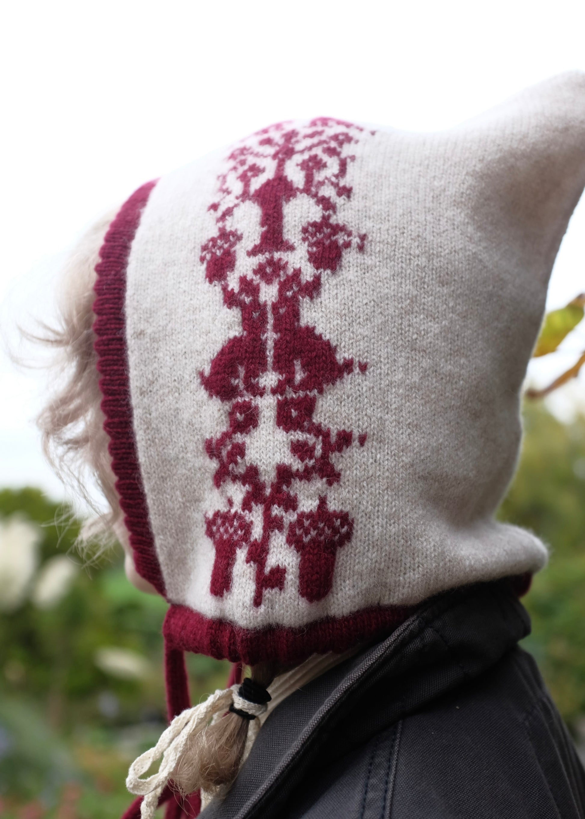 Person wearing a cream knitted bonnet with red floral animal patterns against a blurred natural background