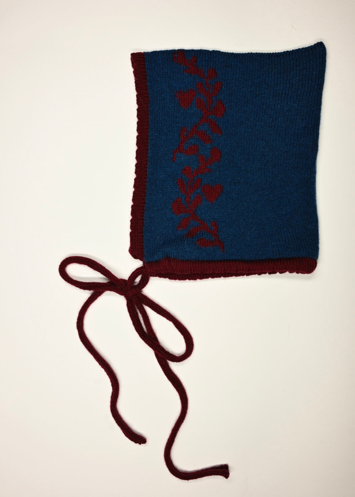 A flat lay of our cashmere knitted bonnet in blue with a red tie, trim and laurel leaf deign 