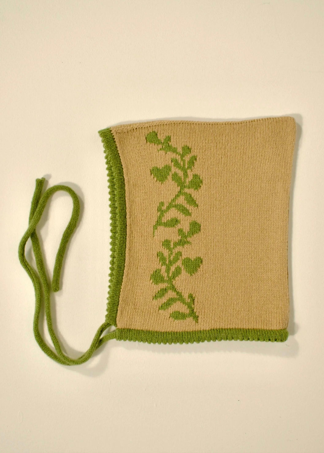 Knitted tan bonnet with green vine laurel leaf pattern on beige background
