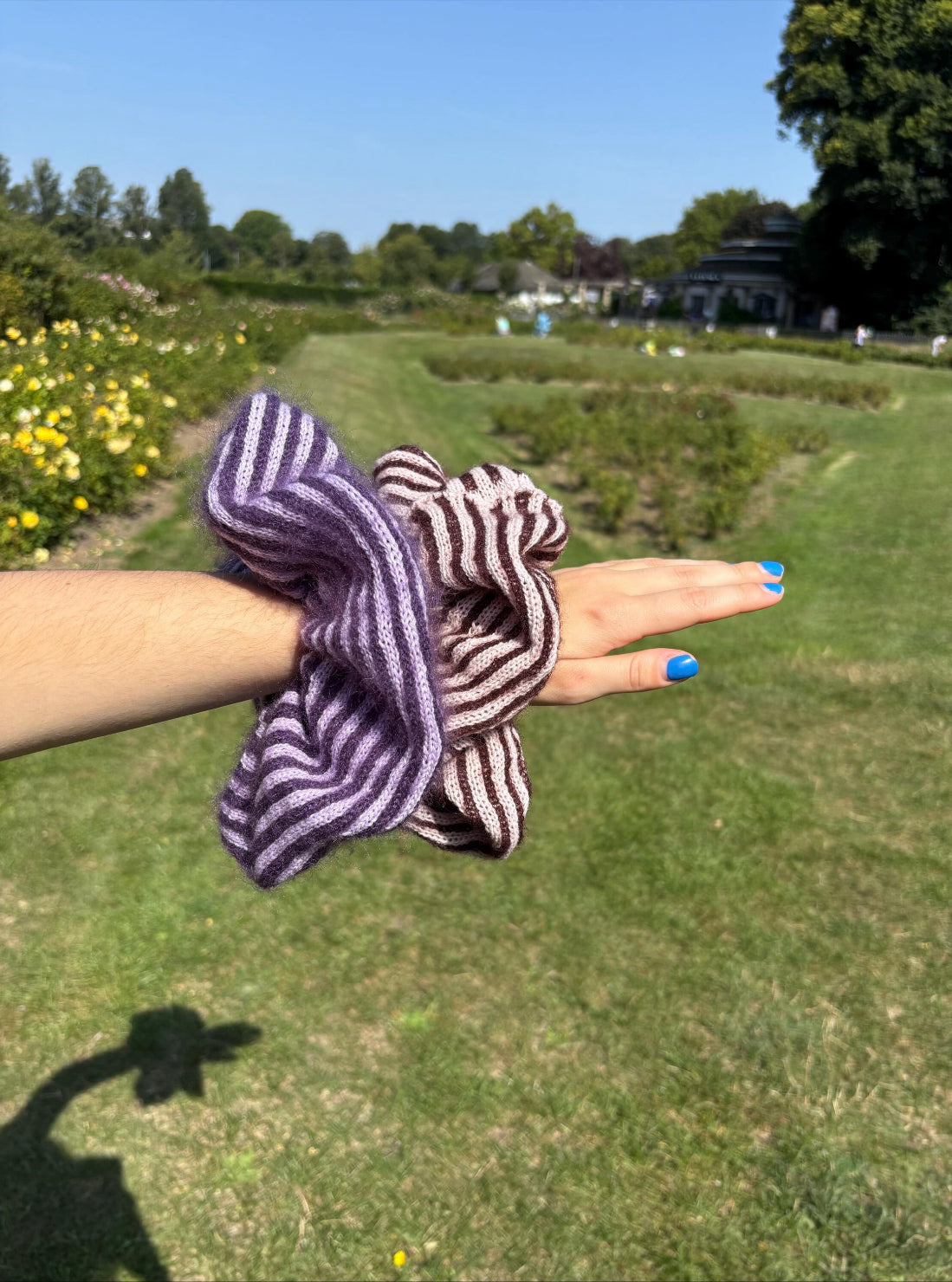 Two striped cashmere and mohair knitted oversized scrunchies displayed on a womens arm with a grassy background demonstrating the colour options, first one being lilac and purple, the second one being plum and pink
