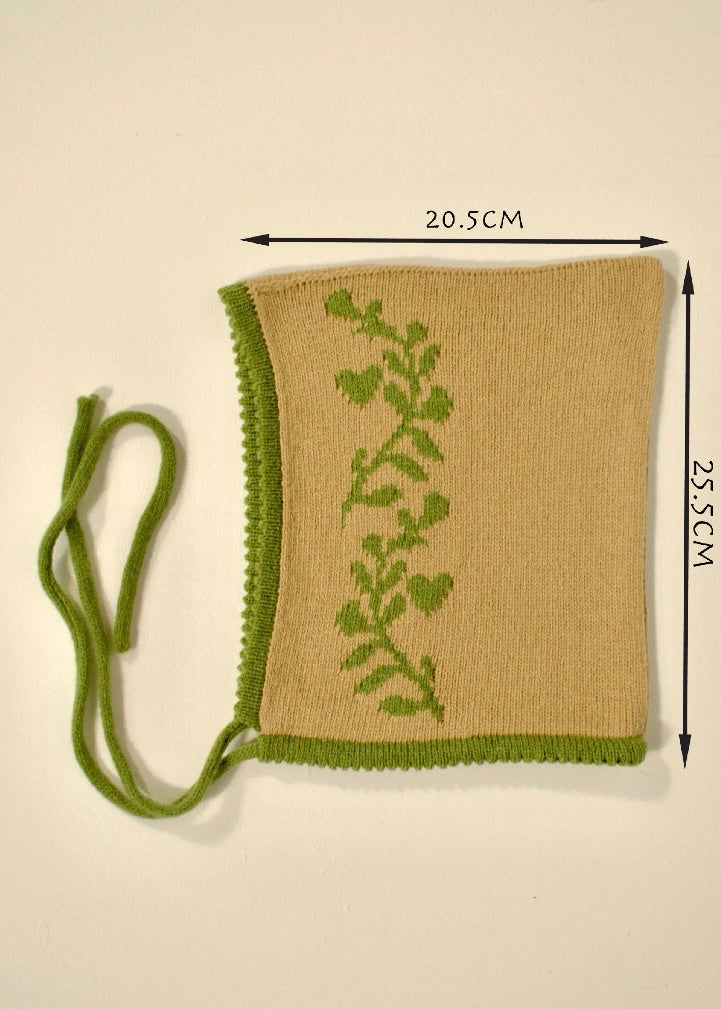 Knitted tan cashmere bonnet with green laurel leaf vine pattern and measurements (20.5cm wide by 25.5cm ) on a beige background