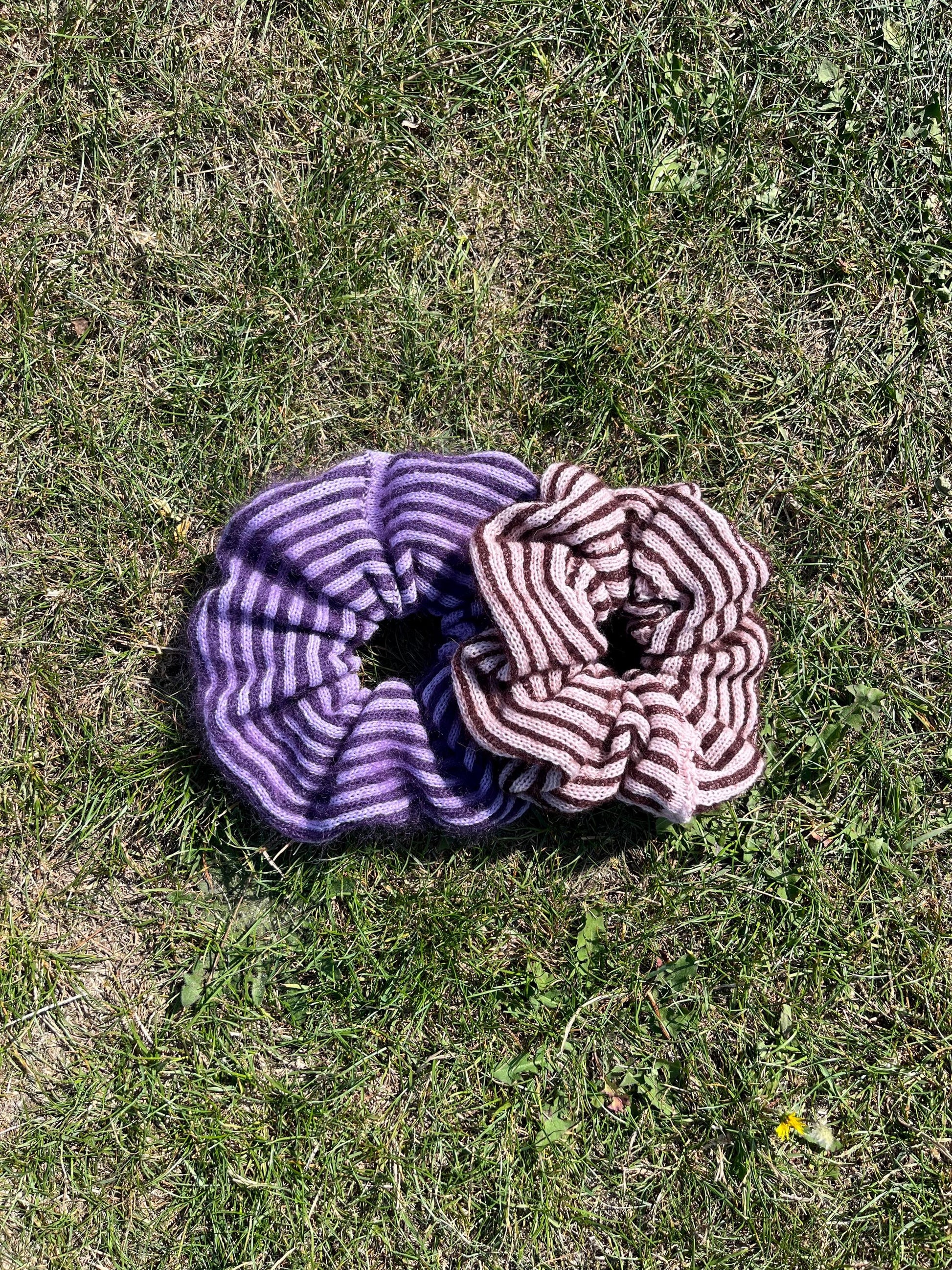 Two striped cashmere and mohair knitted oversized scrunchies lying on grass displaying the colour options, first one being lilac and purple and white and the second one being plum and pink