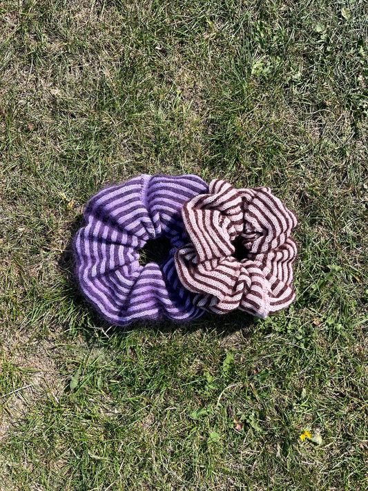 Two striped cashmere and mohair knitted oversized scrunchies lying on grass displaying the colour options, first one being lilac and purple and white and the second one being plum and pink