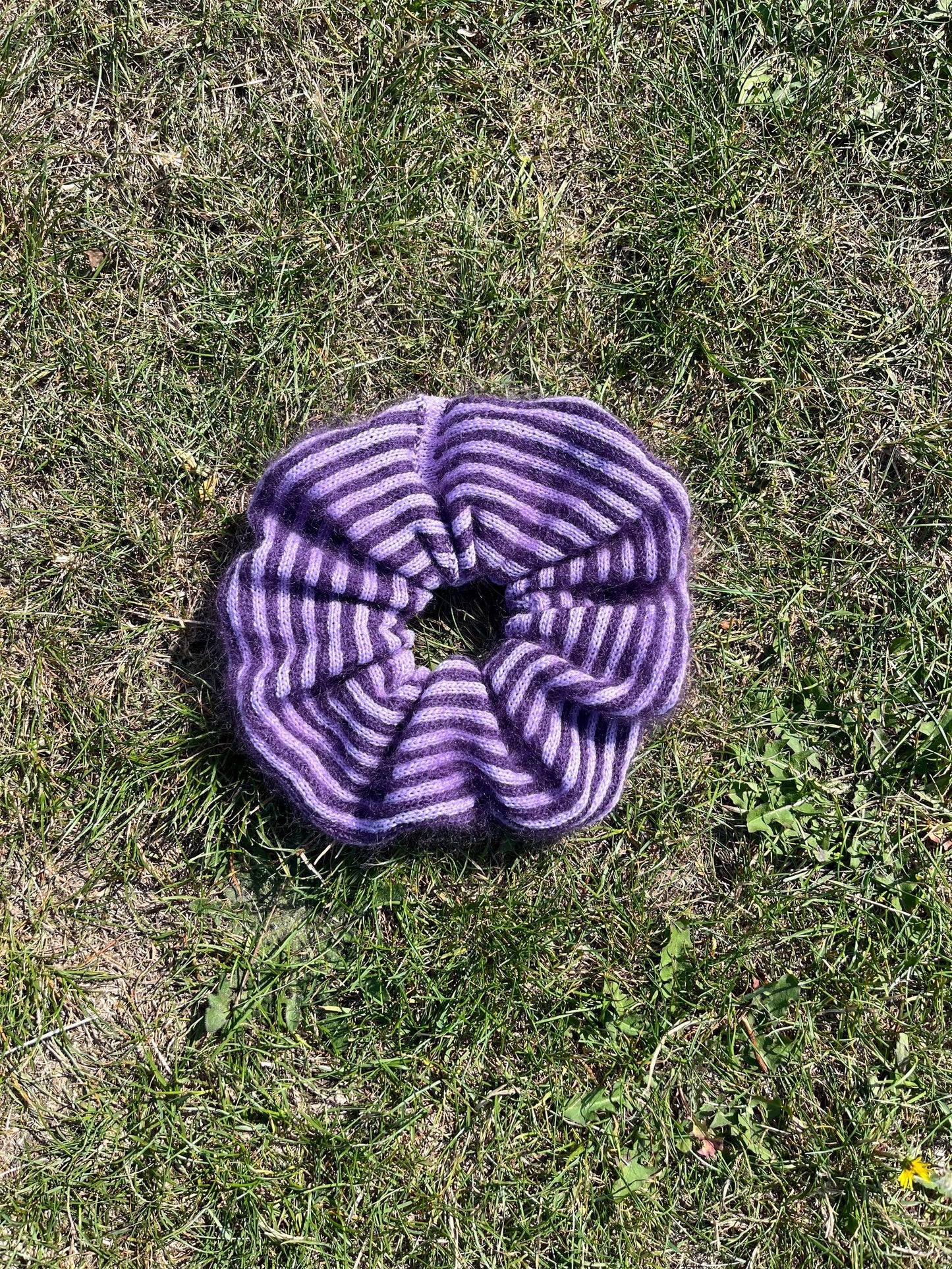 A striped cashmere and mohair knitted oversized scrunchies lying on grass coloured purple and lilac 