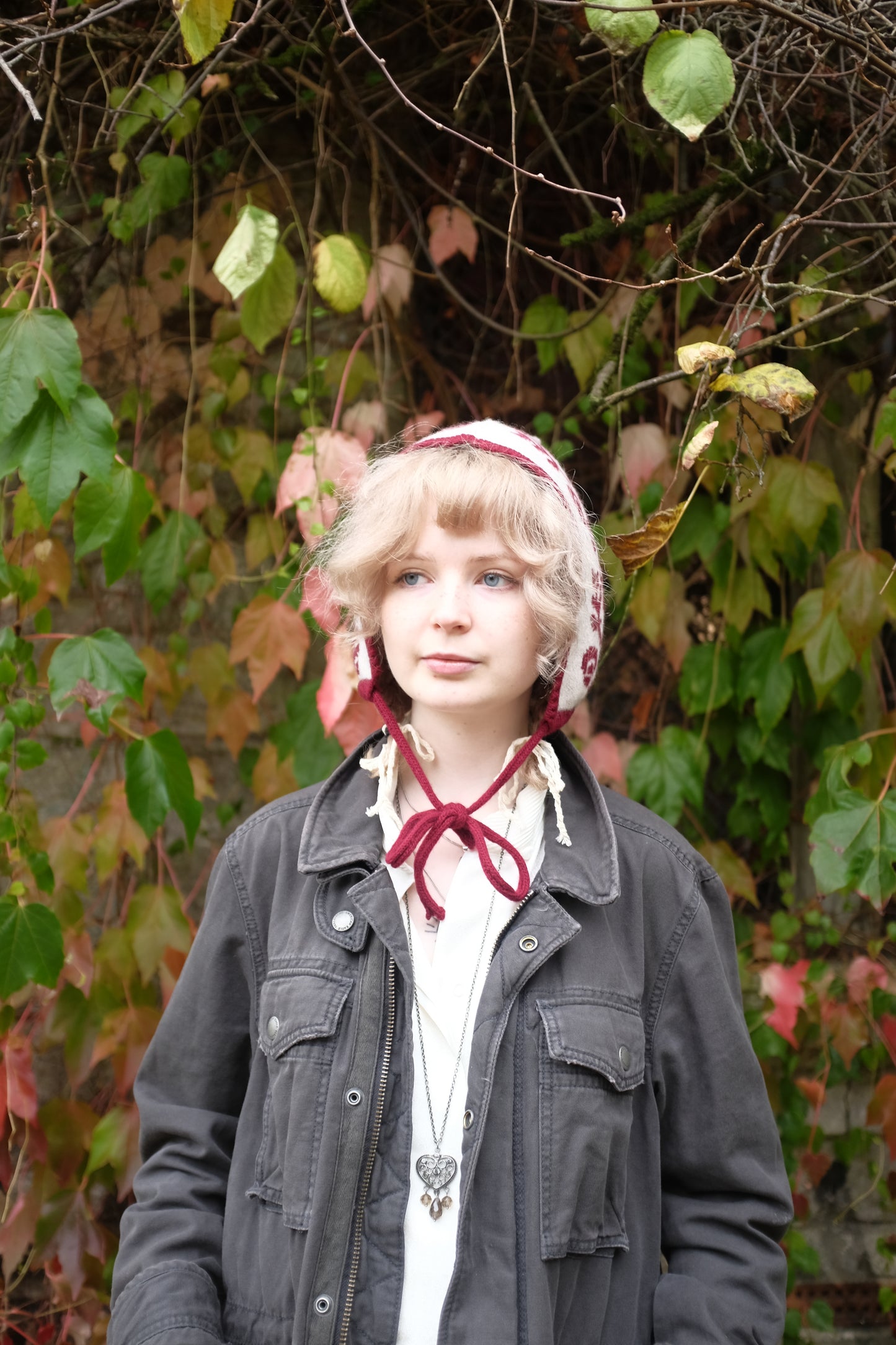 A front view of a women stood looking to the side wearing a cashmere and lambswool knitted bonnet. The base is cream with the trim, pattern and tie being a deep red. The pattern is made up of a range of mushrooms and leaves, scattered satisfyingly up the bonnet. Behind her is a mix of autumnal leaves 