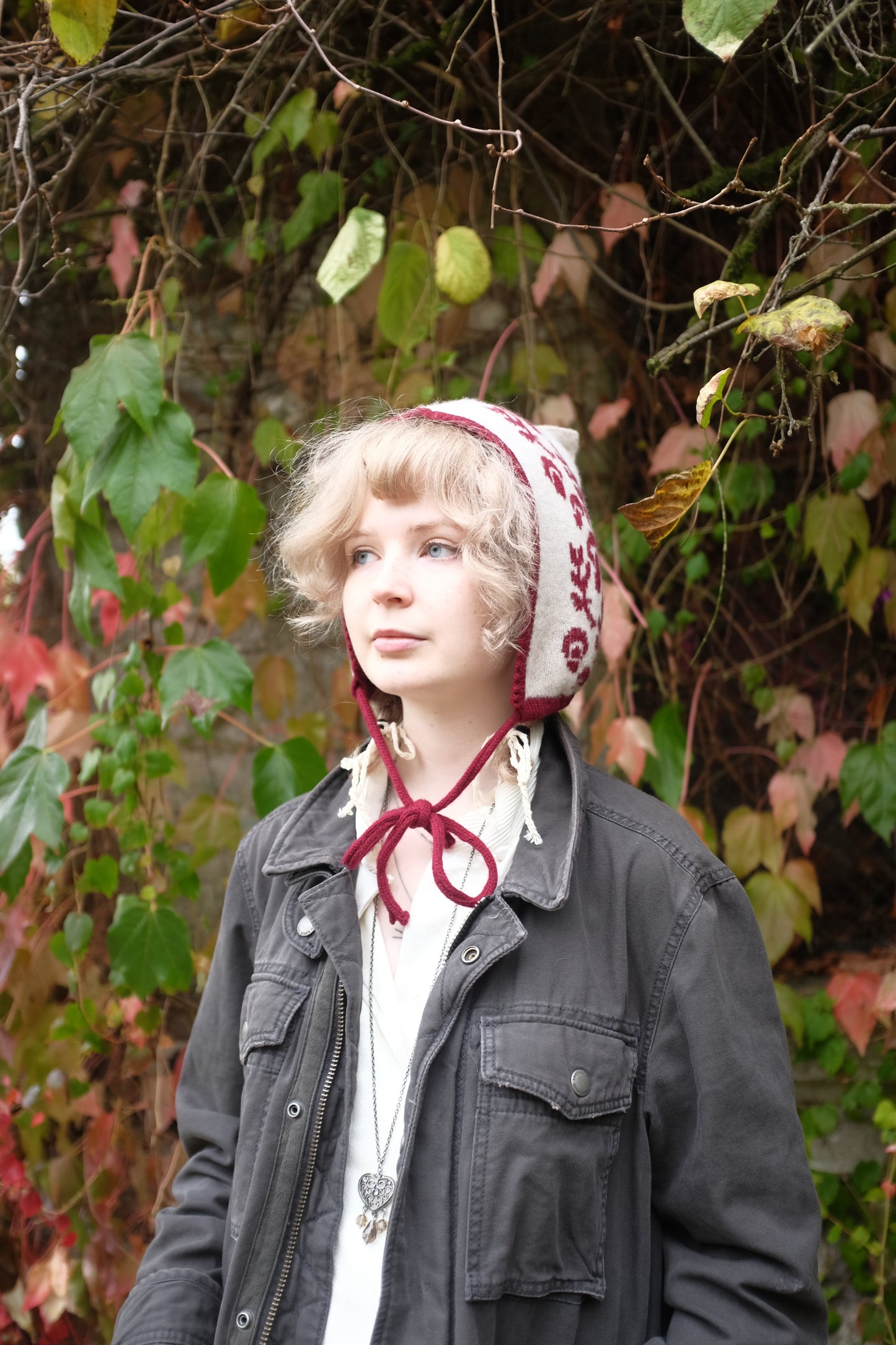 A front view of a women stood looking to the side wearing a cashmere and lambswool knitted bonnet. The base is cream with the trim, pattern and tie being a deep red. The pattern is made up of a range of mushrooms and leaves, scattered satisfyingly up the bonnet. Behind her is a mix of autumnal leaves 