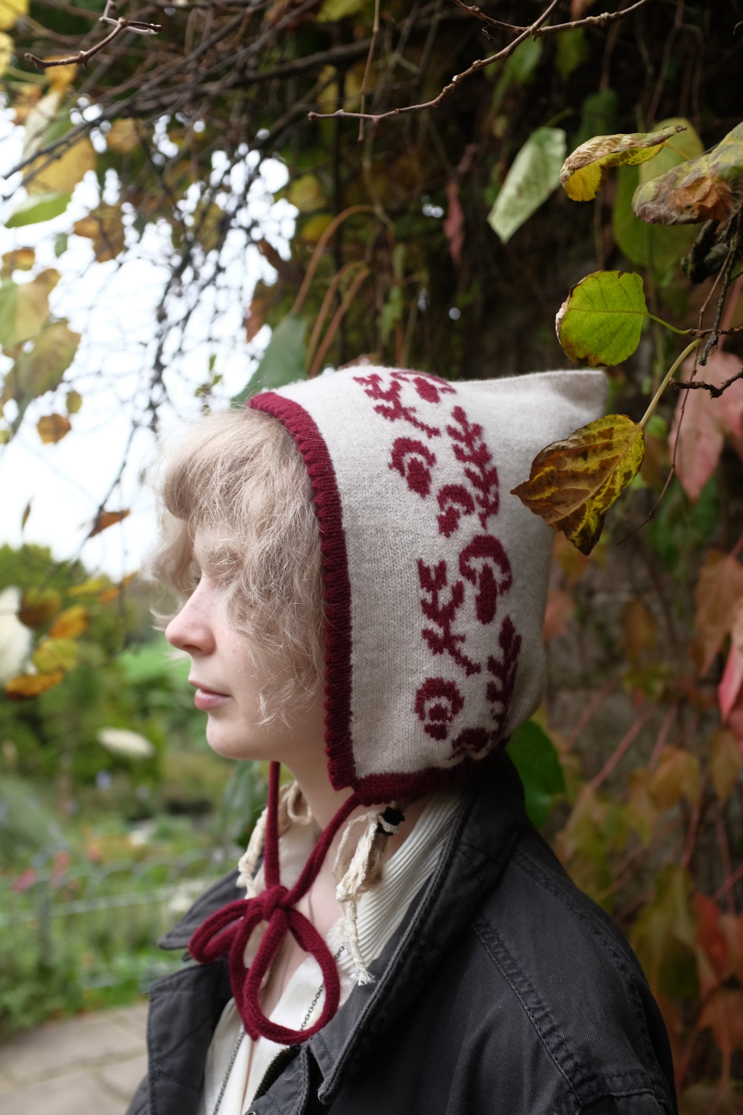 A side view of a women stood looking to the side wearing a cashmere and lambswool knitted bonnet. The base is cream with the trim, pattern and tie being a deep red. The pattern is made up of a range of mushrooms and leaves, scattered satisfyingly up the bonnet. Behind her is a mix of autumnal leaves 