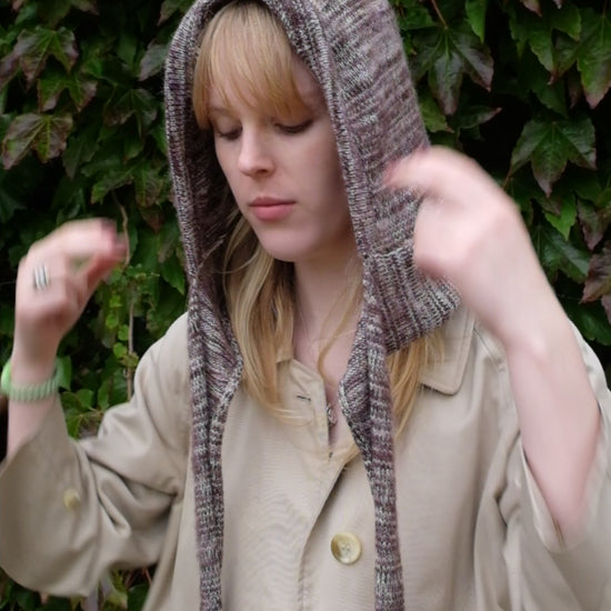 Person wearing a burgundy mohair bonnet-style hood that fully covers the head and neck. With two long, soft extensions hanging down from the front. The hood is thick and textured, offering warmth and a cosy appearance, ideal for cold weather. The person is dressed in a light coloured coat and stands outdoors against a backdrop of green and red foliage 