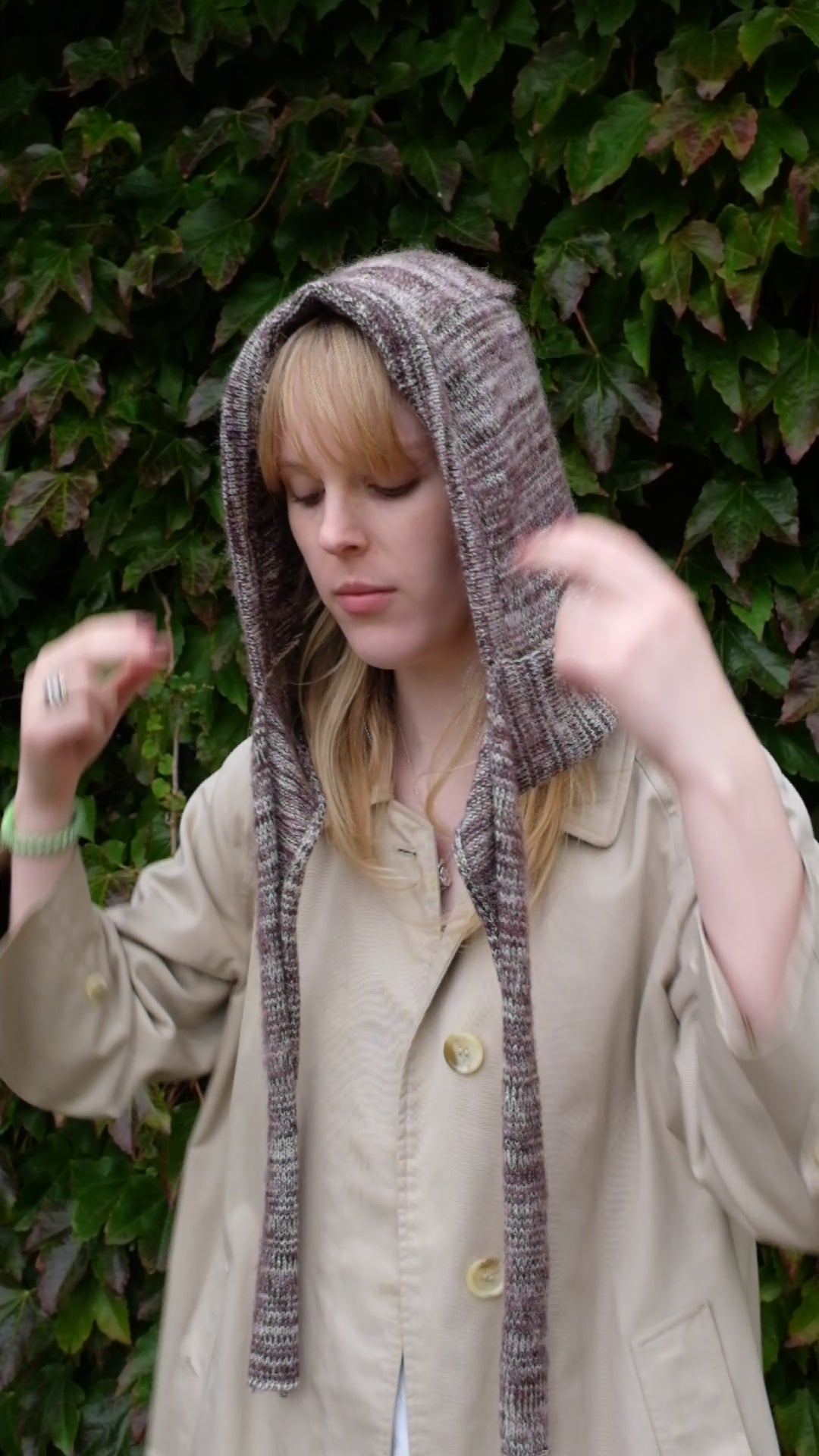 Person wearing a burgundy mohair bonnet-style hood that fully covers the head and neck. With two long, soft extensions hanging down from the front. The hood is thick and textured, offering warmth and a cosy appearance, ideal for cold weather. The person is dressed in a light coloured coat and stands outdoors against a backdrop of green and red foliage 