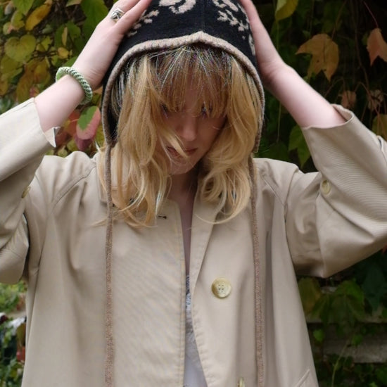 A video of a women tieing a cashmere and lambswool knitted bonnet. The Bonnets base is black whilst the trim, pattern and tie are tan. The pattern on the bonnet consists of leaves and mushrooms sequenced satisfyingly up the side of the hood. Behind the women is a wave of green, red and orange leaves. 