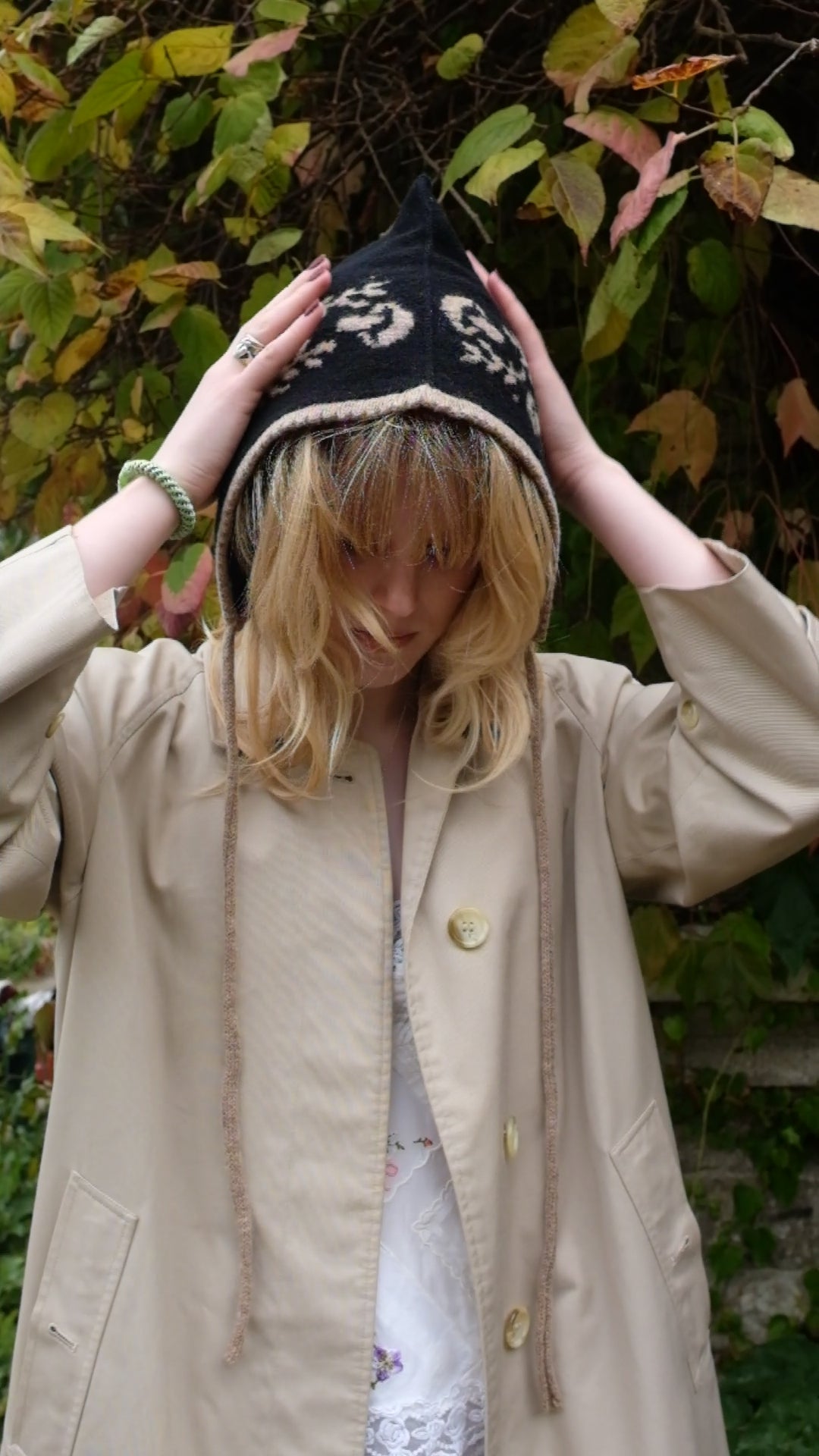 A video of a women tieing a cashmere and lambswool knitted bonnet. The Bonnets base is black whilst the trim, pattern and tie are tan. The pattern on the bonnet consists of leaves and mushrooms sequenced satisfyingly up the side of the hood. Behind the women is a wave of green, red and orange leaves. 
