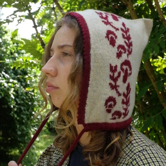 A women from a side angel, videoed tieing a cashmere and lambswool knitted bonnet. The base is cream with the trim, pattern and tie being a deep red. The pattern is made up a range of mushrooms leaves, scattered satisfyingly up the bonnet. Behind her are large green trees highlighted by sunlight 