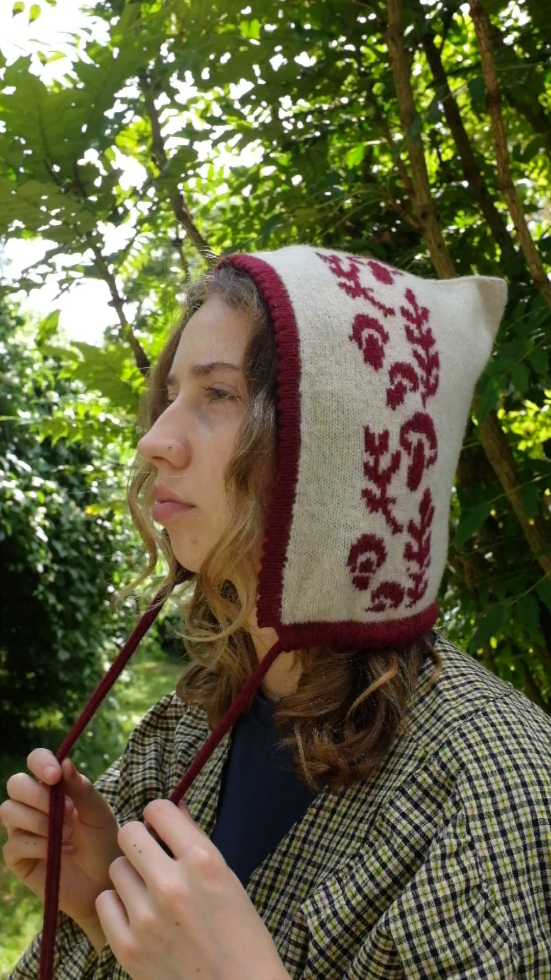 A women from a side angel, videoed tieing a cashmere and lambswool knitted bonnet. The base is cream with the trim, pattern and tie being a deep red. The pattern is made up a range of mushrooms leaves, scattered satisfyingly up the bonnet. Behind her are large green trees highlighted by sunlight 