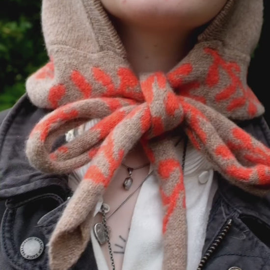 A video of a woman wearing a lambswool knitted bonnet hood. The base of the bonnet is a tan colour. There is a fire orange pattern on the bottom of the bonnet tie that vis longer, mimicking a scarf. The pattern on the is a sequenced of laurel leaves and hearts. The video itself shows the versatility of the bonnet, firstly the tie being tied in a bow, and secondly it draped around her neck and shoulder. 