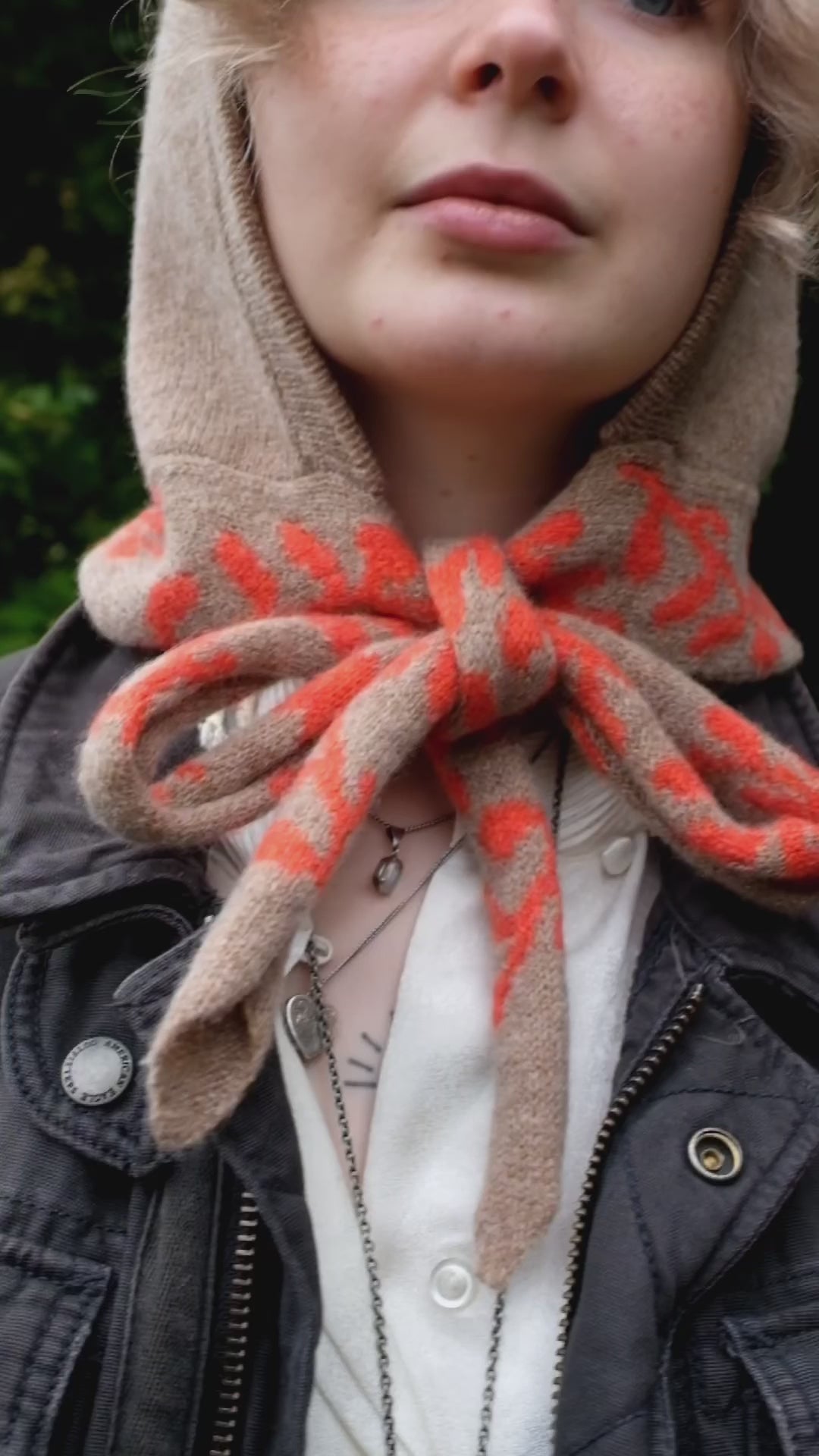 A video of a woman wearing a lambswool knitted bonnet hood. The base of the bonnet is a tan colour. There is a fire orange pattern on the bottom of the bonnet tie that vis longer, mimicking a scarf. The pattern on the is a sequenced of laurel leaves and hearts. The video itself shows the versatility of the bonnet, firstly the tie being tied in a bow, and secondly it draped around her neck and shoulder. 