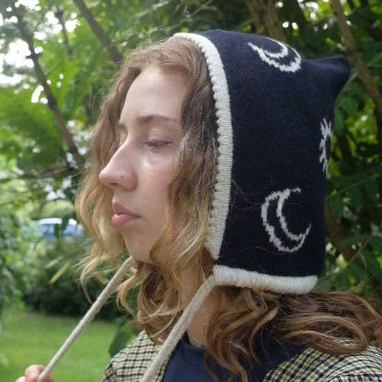 A video from a side angled view A female model wearing a knitted bonnet with a navy base and cream and gold moon and sun pattern. It has a cream and gold trim around the face with a drawstring. The video is a closer up view showing the detail of the moon and sun pattern 