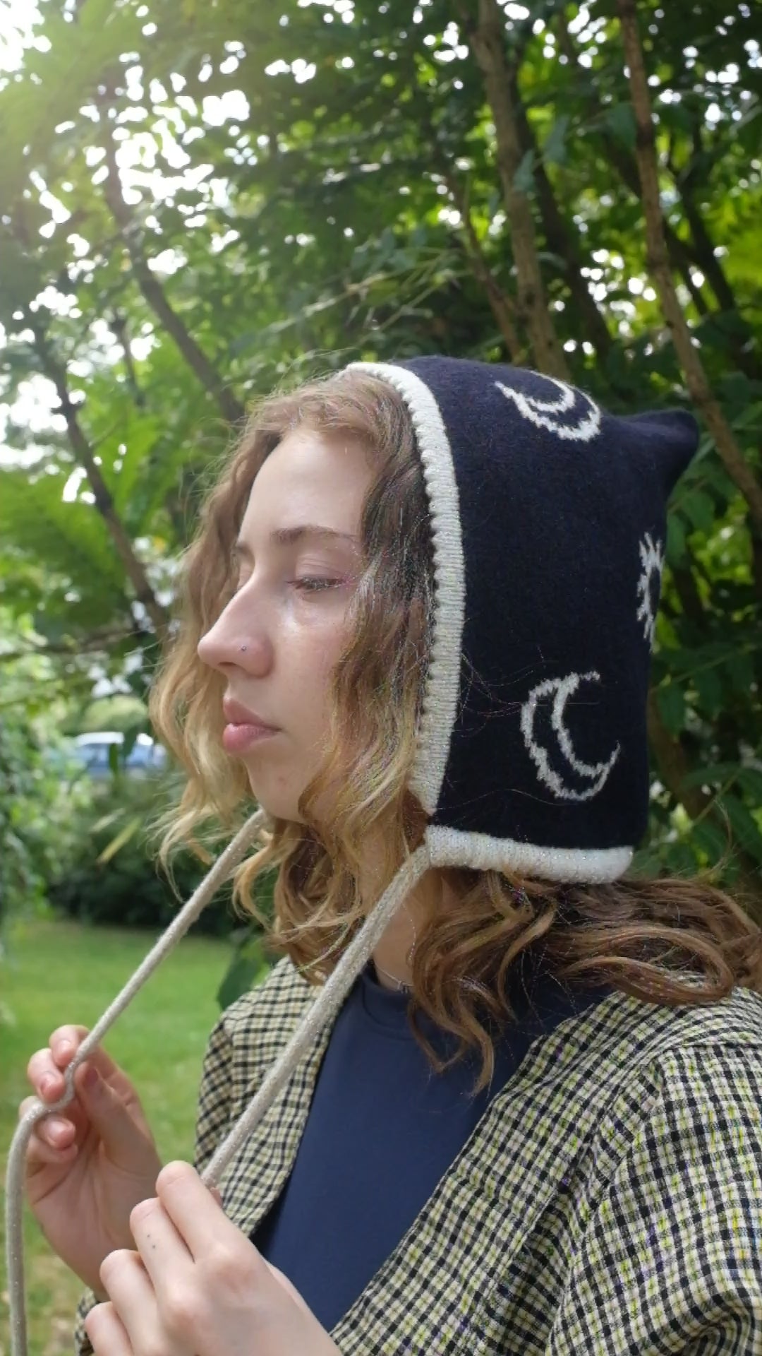 A video from a side angled view A female model wearing a knitted bonnet with a navy base and cream and gold moon and sun pattern. It has a cream and gold trim around the face with a drawstring. The video is a closer up view showing the detail of the moon and sun pattern 
