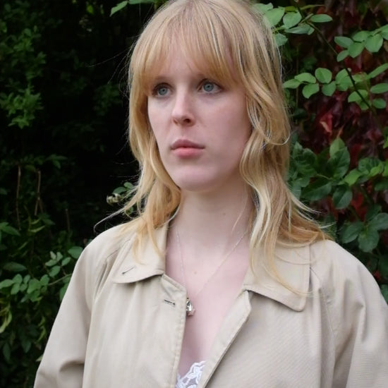 A video of a women wearing a lambswool hand crafted bonnet, with a scarf detailing around the bottom. The main colour of the bonnet is navy. With a white trim, and around the scarf detailing there is a celestial print of the moon and stars in white with gold shimmering details. Behind the women is a blurry mix of green and red leaves 