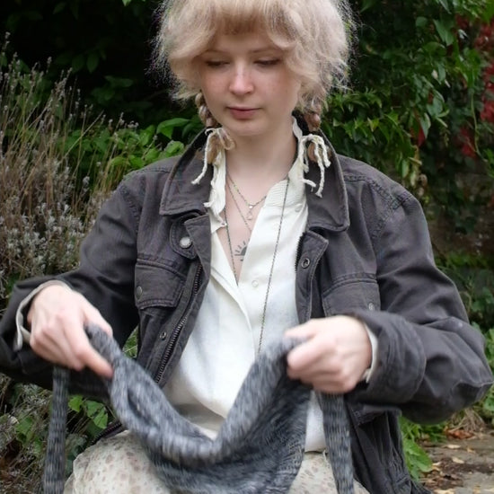 Person wearing a black mohair bonnet-style hood that fully covers the head and neck. With two long, soft extensions hanging down from the front. The hood is thick and textured, offering warmth and a cosy appearance, ideal for cold weather. The person is dressed in a light coloured coat and stands outdoors against a backdrop of green and red foliage 